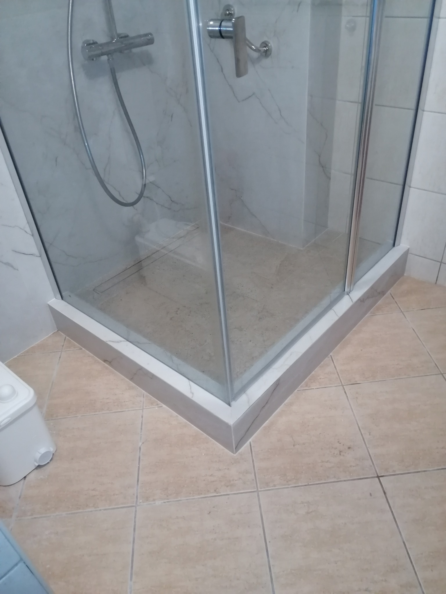 Nowoczesna kabina prysznicowa z marmurowym wykończeniem, widok z dołu, ukazujący detale płytek i brodzika. Chromowana armatura i szklane drzwi.