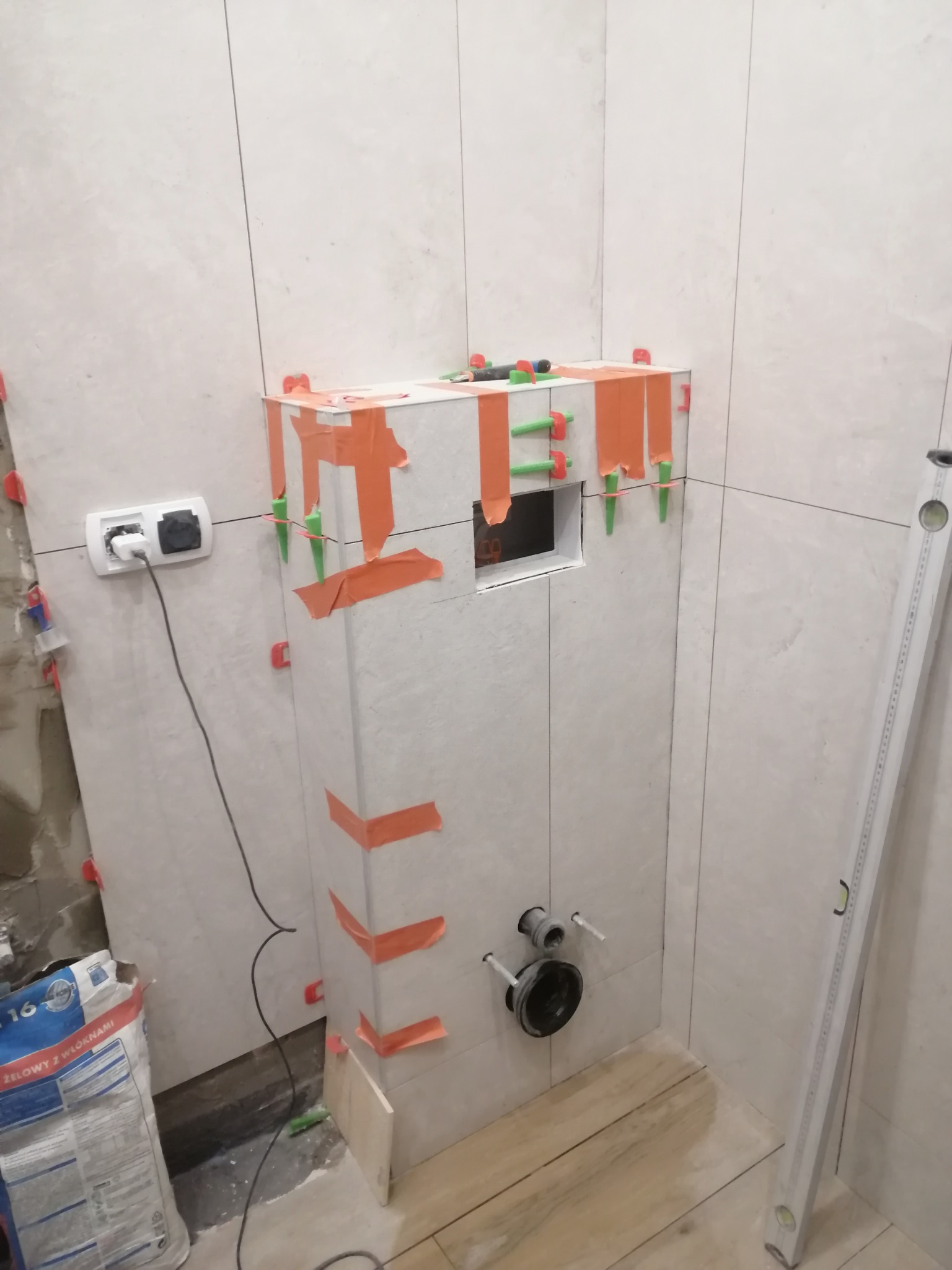 Białe płytki na zabudowie stelaża WC w trakcie montażu. Widoczne poziomica, taśma malarska i krzyżyki glazurnicze. W tle ściana z gniazdkiem elektrycznym.