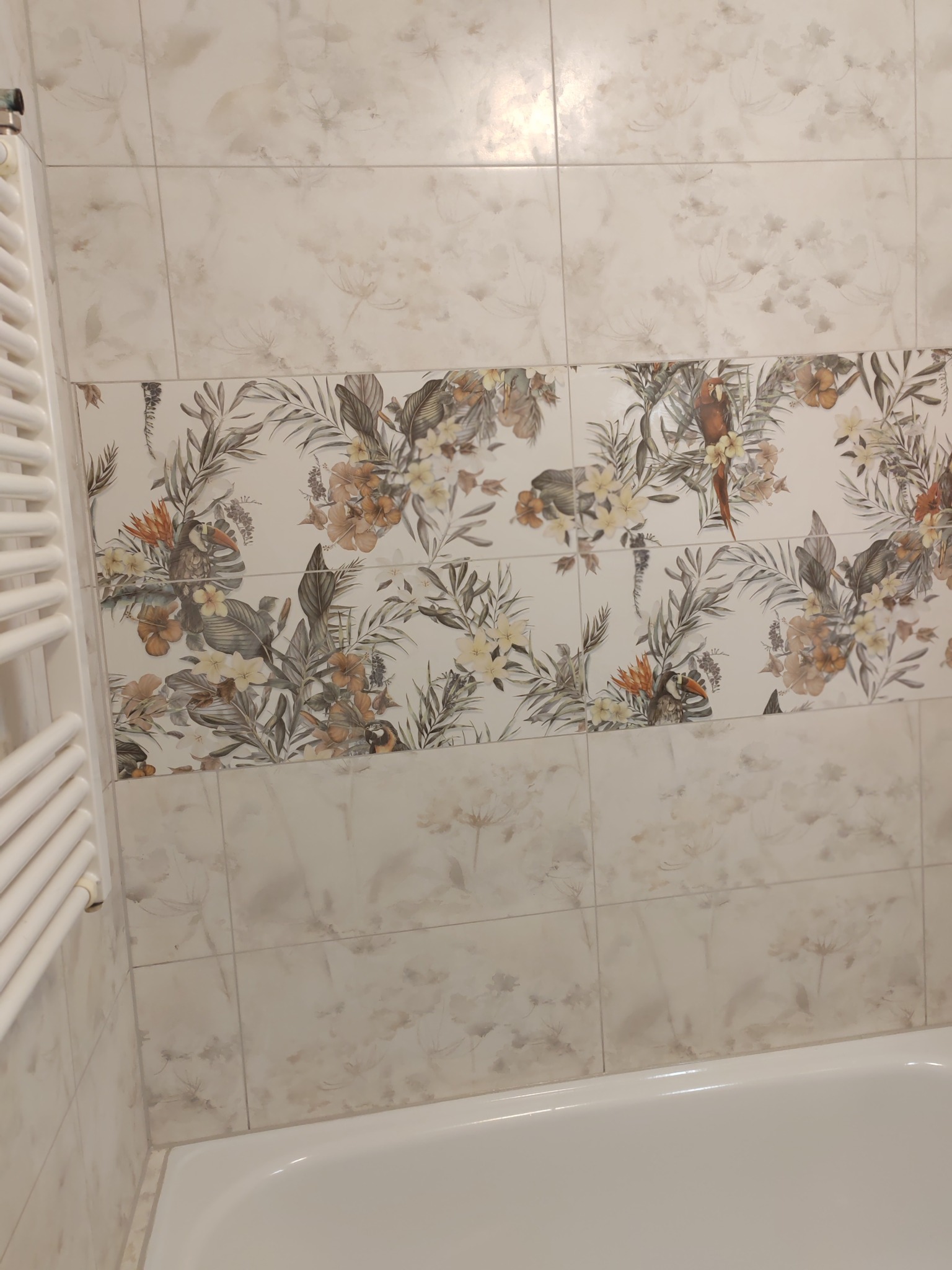 Wykończona ściana łazienki z beżowymi płytkami w kwiaty i papugi, nad białą wanną. Widoczny biały grzejnik drabinkowy.