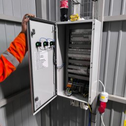 ANDRZEJ JÓŹWIAK FIRMA HANDLOWO-USŁUGOWA Black Car - Otwarta skrzynka elektryczna z listwą zaciskową i opisem obwodów, widoczna ręka elektryka w pomarańczowym ubraniu roboczym. Instalacja w szarej hali.