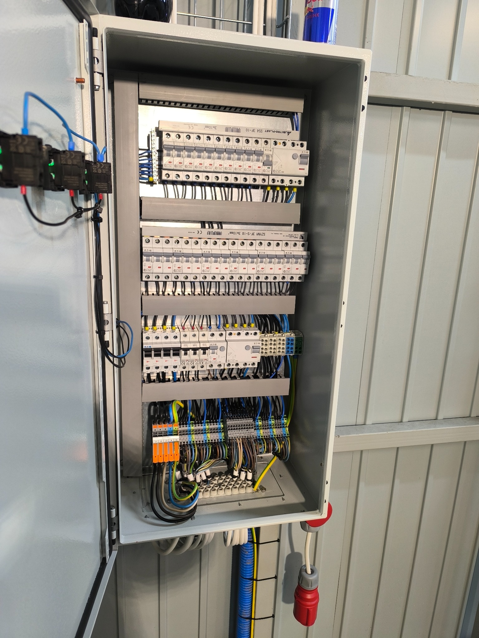 Otwarta skrzynka elektryczna z bezpiecznikami i okablowaniem wewnątrz, widoczne elementy instalacji elektrycznej, kable w różnych kolorach, szyny DIN i aparatura modułowa.