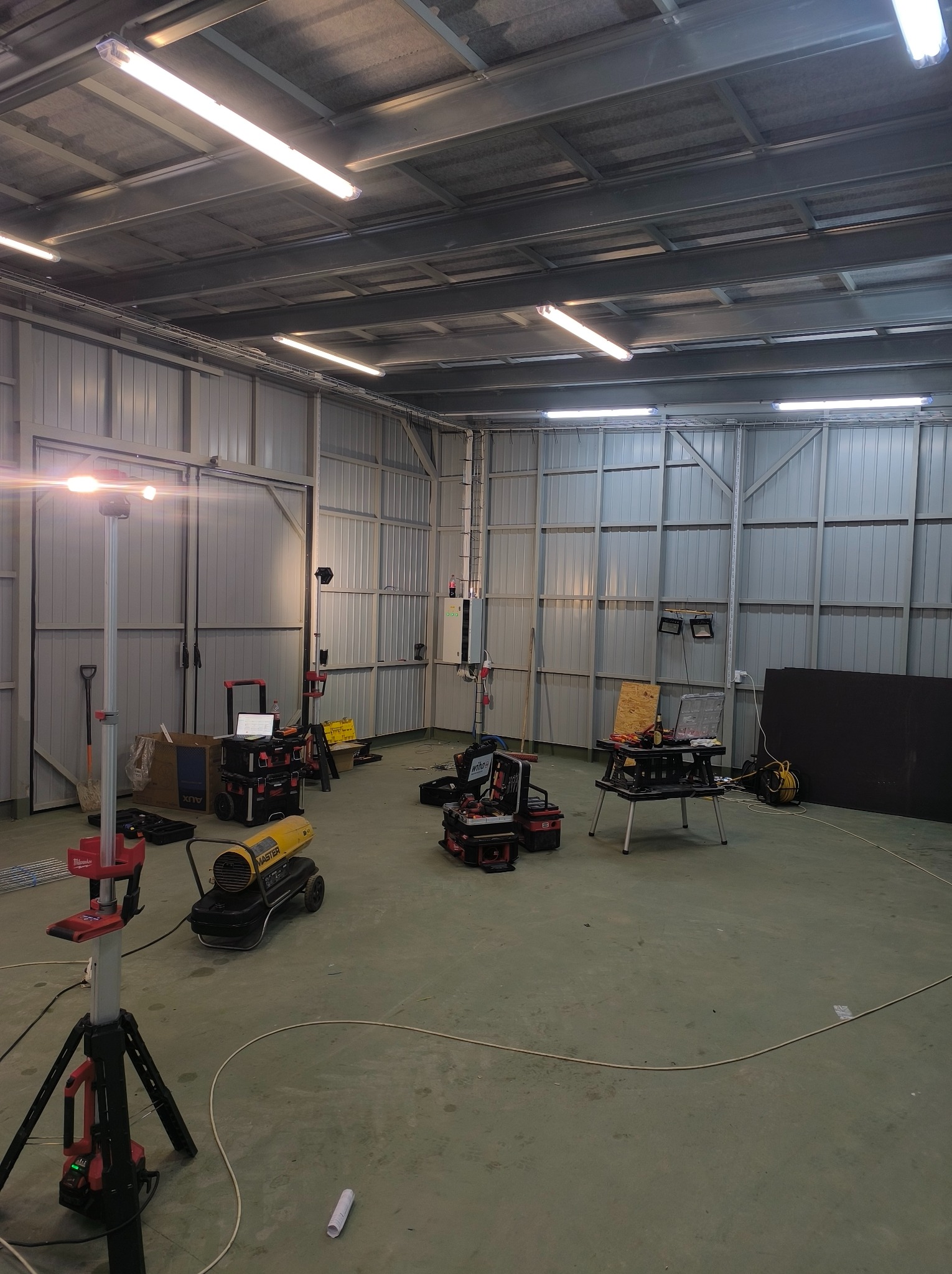 Wnętrze hali z metalowymi ścianami i sufitami, oświetlone lampami. Widoczne narzędzia, skrzynki narzędziowe, nagrzewnica i lampa na statywie. Przygotowanie do prac.