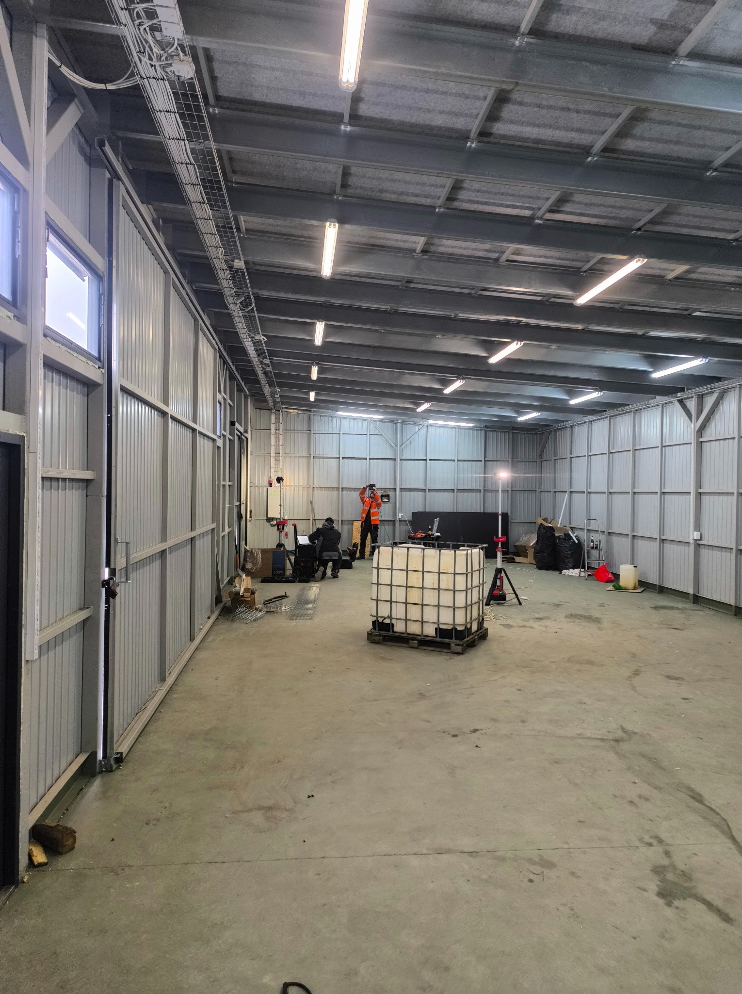 Wnętrze hali z metalową konstrukcją, gdzie ekipa w kamizelkach montuje instalacje pod oświetlenie. Widoczne kable w korytkach i lampy. Hala w trakcie prac.