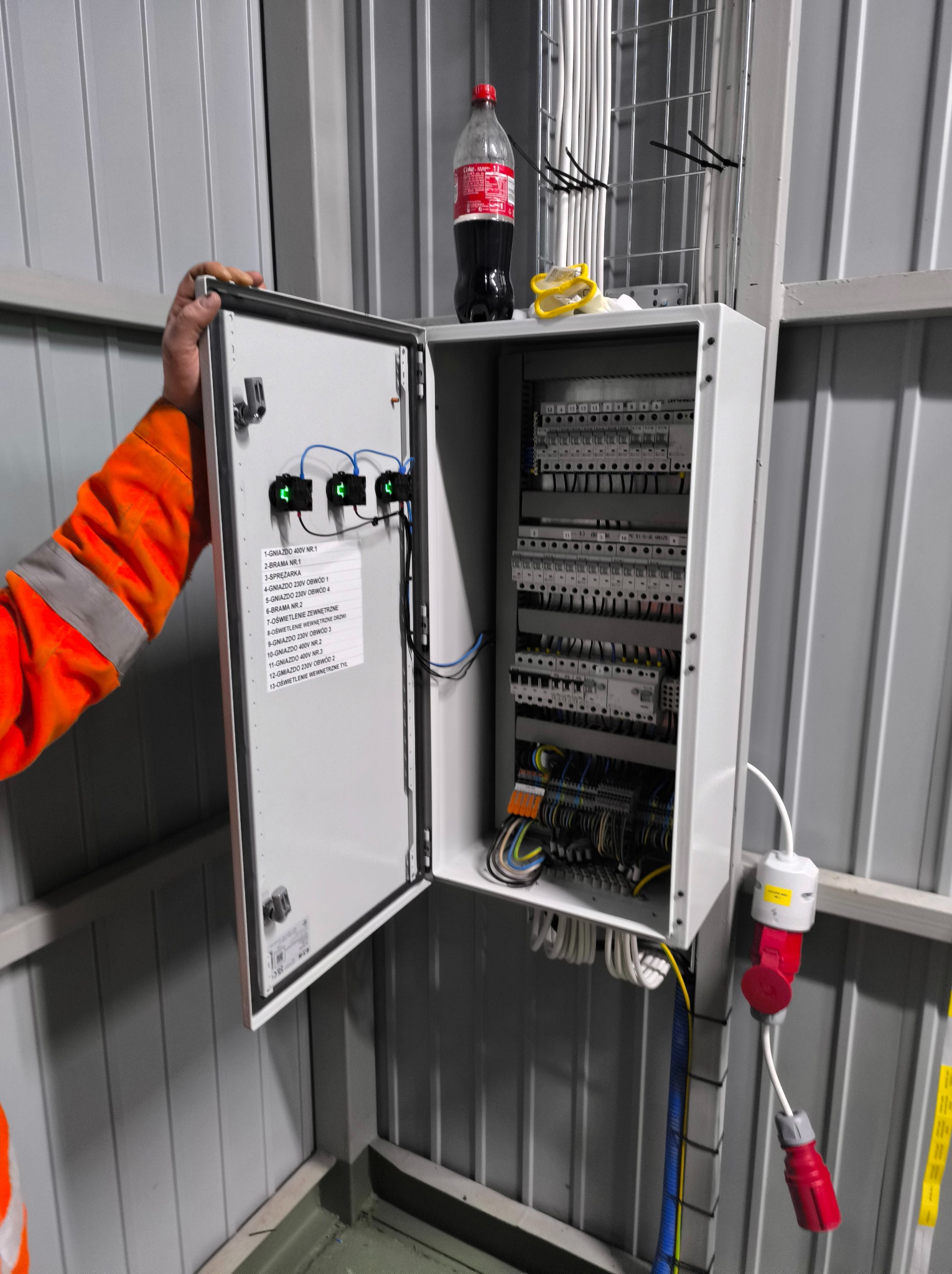 Otwarta skrzynka elektryczna z listwą zaciskową i opisem obwodów, widoczna ręka elektryka w pomarańczowym ubraniu roboczym. Instalacja w szarej hali.
