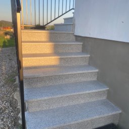 USŁUGI KAMIENIARSKO-FLIZIARSKIE Józef Sukiennik - Zewnętrzne schody z jasnoszarego kamienia z czarną, metalową balustradą, prowadzące do budynku z jasną elewacją. Widok z dołu, w tle zieleń i błękitne niebo.
