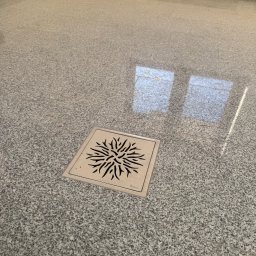 USŁUGI KAMIENIARSKO-FLIZIARSKIE Józef Sukiennik - Szara, polerowana posadzka z kamienia naturalnego z odpływem liniowym w kształcie kwadratu z dekoracyjnym wzorem. Widoczne refleksy światła na powierzchni.