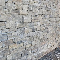 USŁUGI KAMIENIARSKO-FLIZIARSKIE Józef Sukiennik - Ściana z nieregularnego kamienia w odcieniach szarości i beżu, ułożona z widocznymi spoinami. U dołu widoczne resztki gruzu i poziomica.