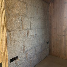 USŁUGI KAMIENIARSKO-FLIZIARSKIE Józef Sukiennik - Ściana wykończona szarym kamieniem w regularne prostokąty, z widocznymi puszkami elektrycznymi i drewnianą boazerią na sąsiedniej ścianie. Surowy stan wykończenia.