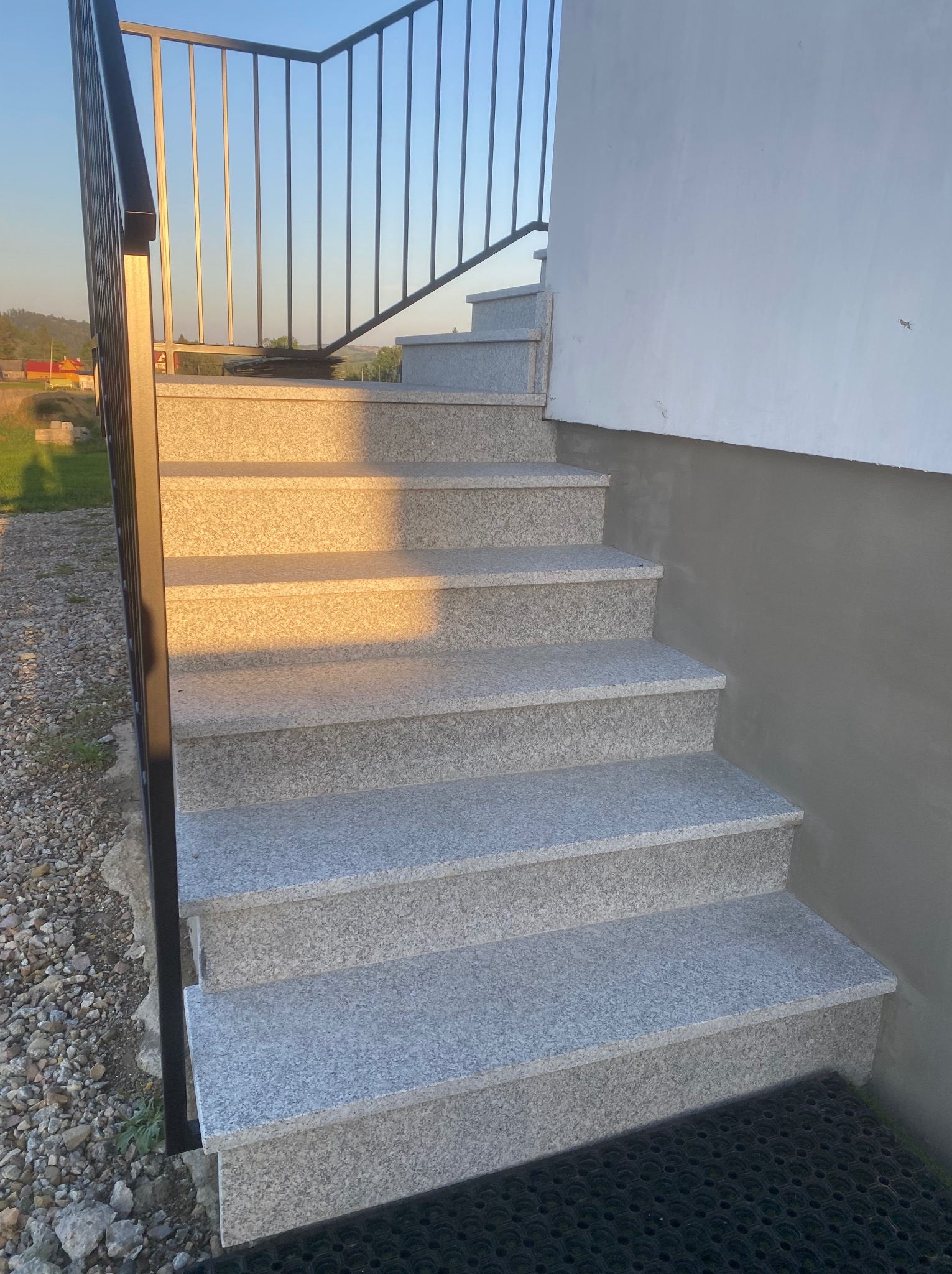 Zewnętrzne schody z jasnoszarego kamienia z czarną, metalową balustradą, prowadzące do budynku z jasną elewacją. Widok z dołu, w tle zieleń i błękitne niebo.