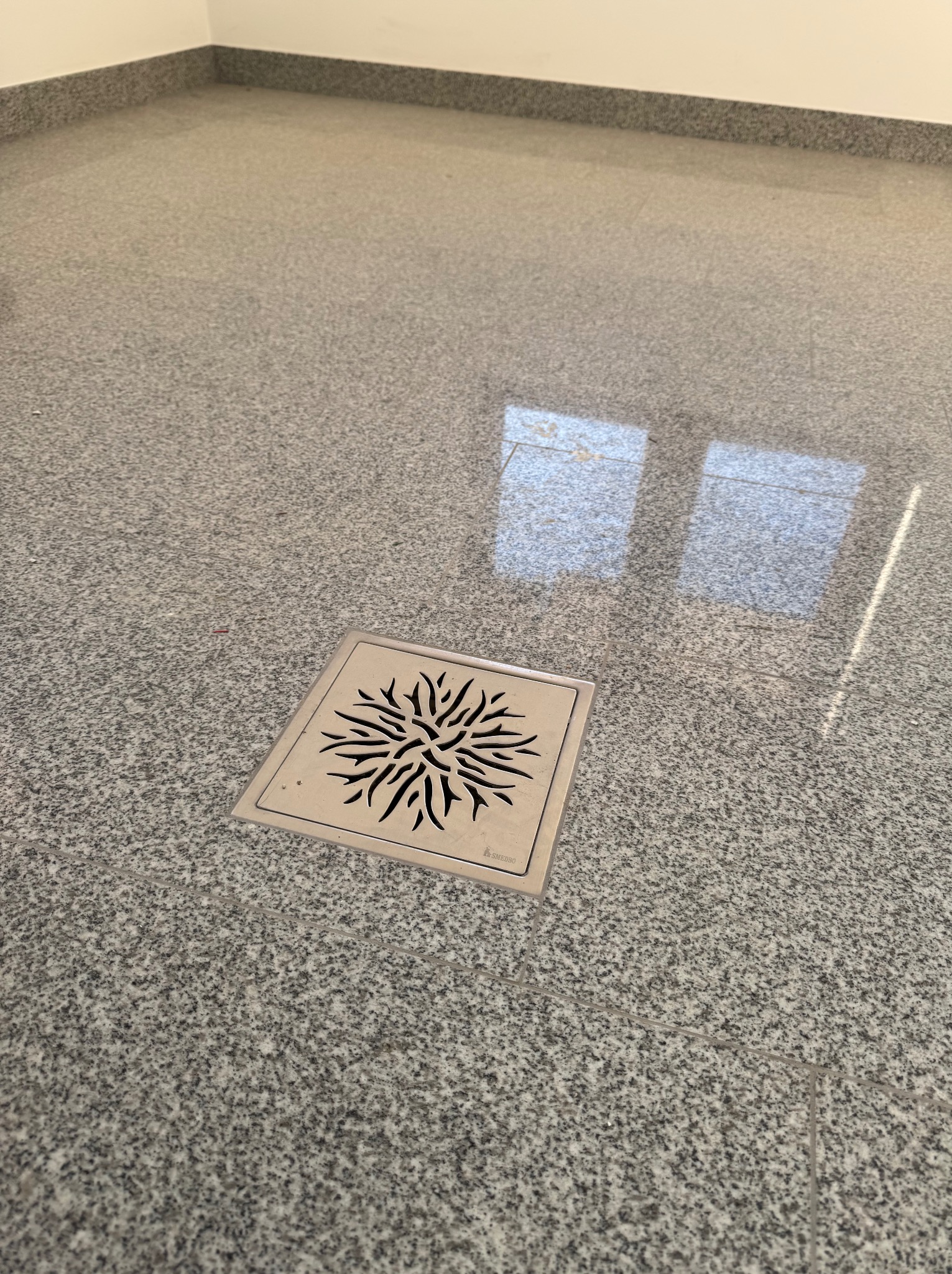 Szara, polerowana posadzka z kamienia naturalnego z odpływem liniowym w kształcie kwadratu z dekoracyjnym wzorem. Widoczne refleksy światła na powierzchni.