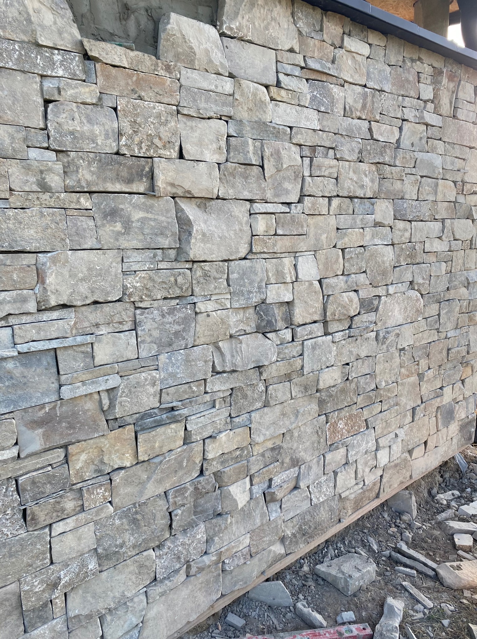 Ściana z nieregularnego kamienia w odcieniach szarości i beżu, ułożona z widocznymi spoinami. U dołu widoczne resztki gruzu i poziomica.