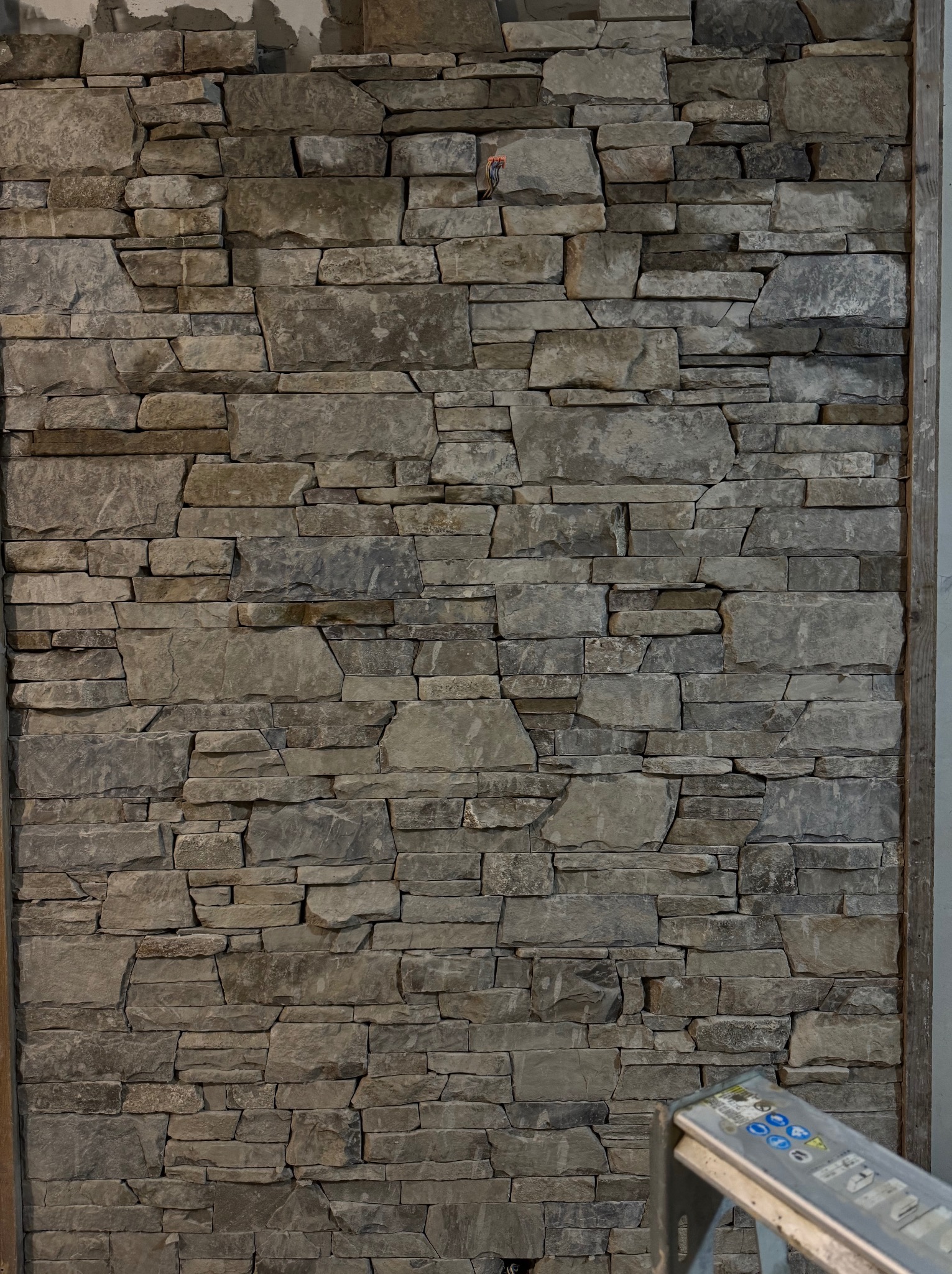 Ściana wykończona nieregularnym kamieniem w odcieniach szarości i beżu, z widocznym fragmentem drabiny w prawym dolnym rogu. Surowe wykończenie, naturalna tekstura.