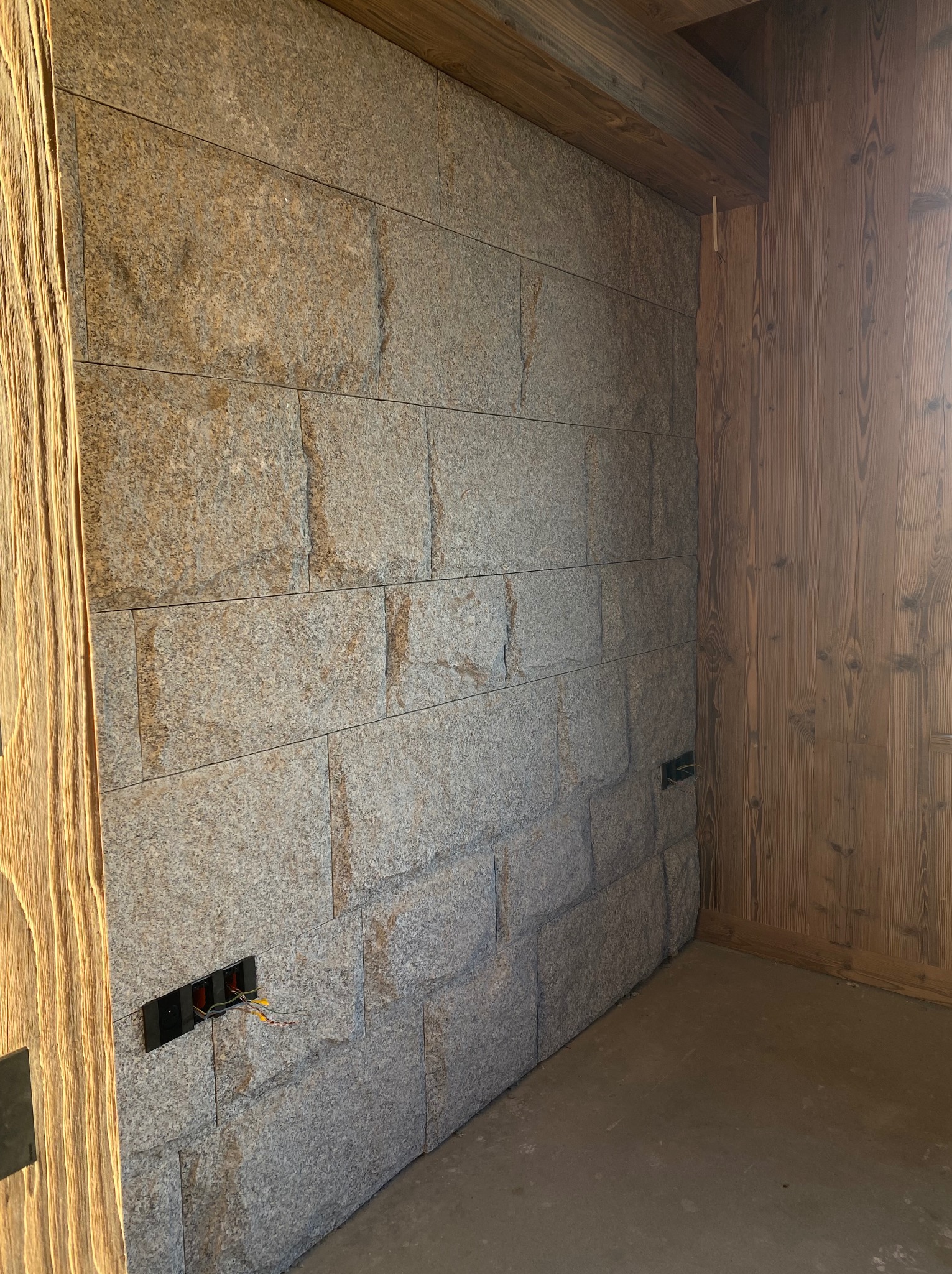 Ściana wykończona szarym kamieniem w regularne prostokąty, z widocznymi puszkami elektrycznymi i drewnianą boazerią na sąsiedniej ścianie. Surowy stan wykończenia.
