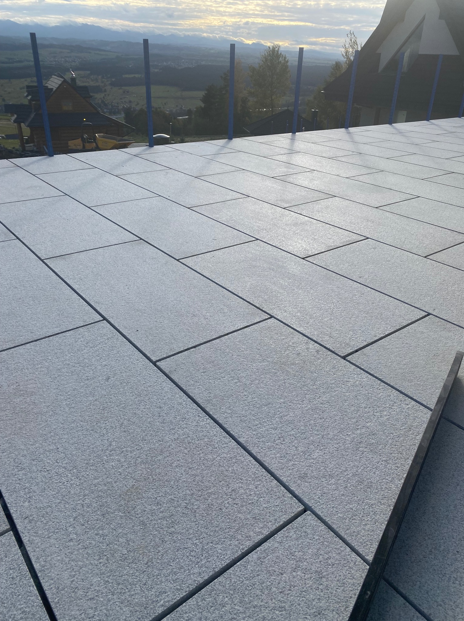 Nowy taras z szarych, prostokątnych płyt kamiennych, ułożonych w regularny wzór. W tle widok na góry i zabudowania. Jedna płyta kamienna jest podniesiona.
