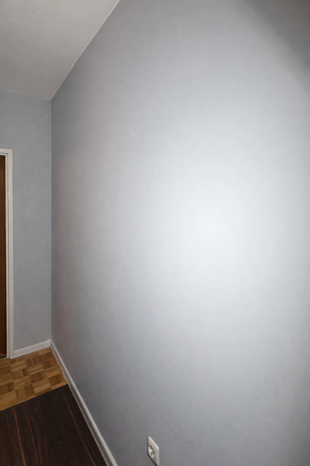 Świeżo pomalowana, gładka ściana w odcieniach szarości w rogu pokoju, widoczny fragment parkietu i gniazdko elektryczne. Minimalistyczne wykończenie wnętrza.