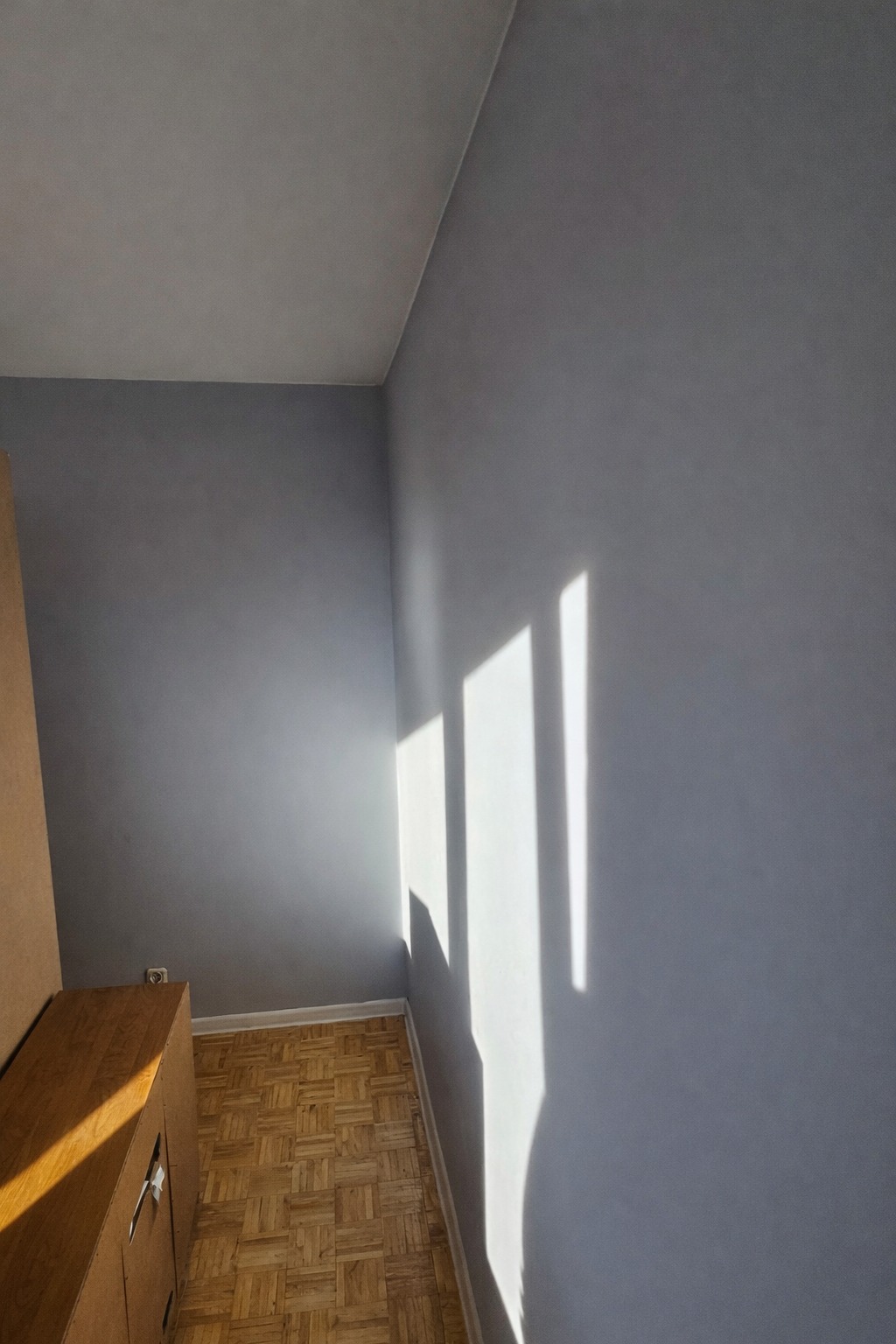 Świeżo pomalowana szara ściana w pokoju z parkietem, oświetlona promieniami słońca tworzącymi geometryczne wzory. Widoczny fragment drewnianej szafki.