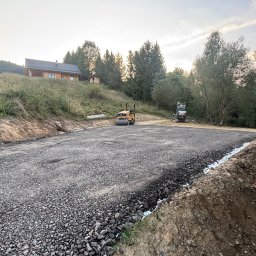 Łopiennik Usługi Turystyczne i Budowlane Mariusz Kaczmar - Utwardzony teren pod budowę w górskim krajobrazie. Walec drogowy i minikoparka na przygotowanym placu. W tle drewniany dom i zalesione wzgórza.