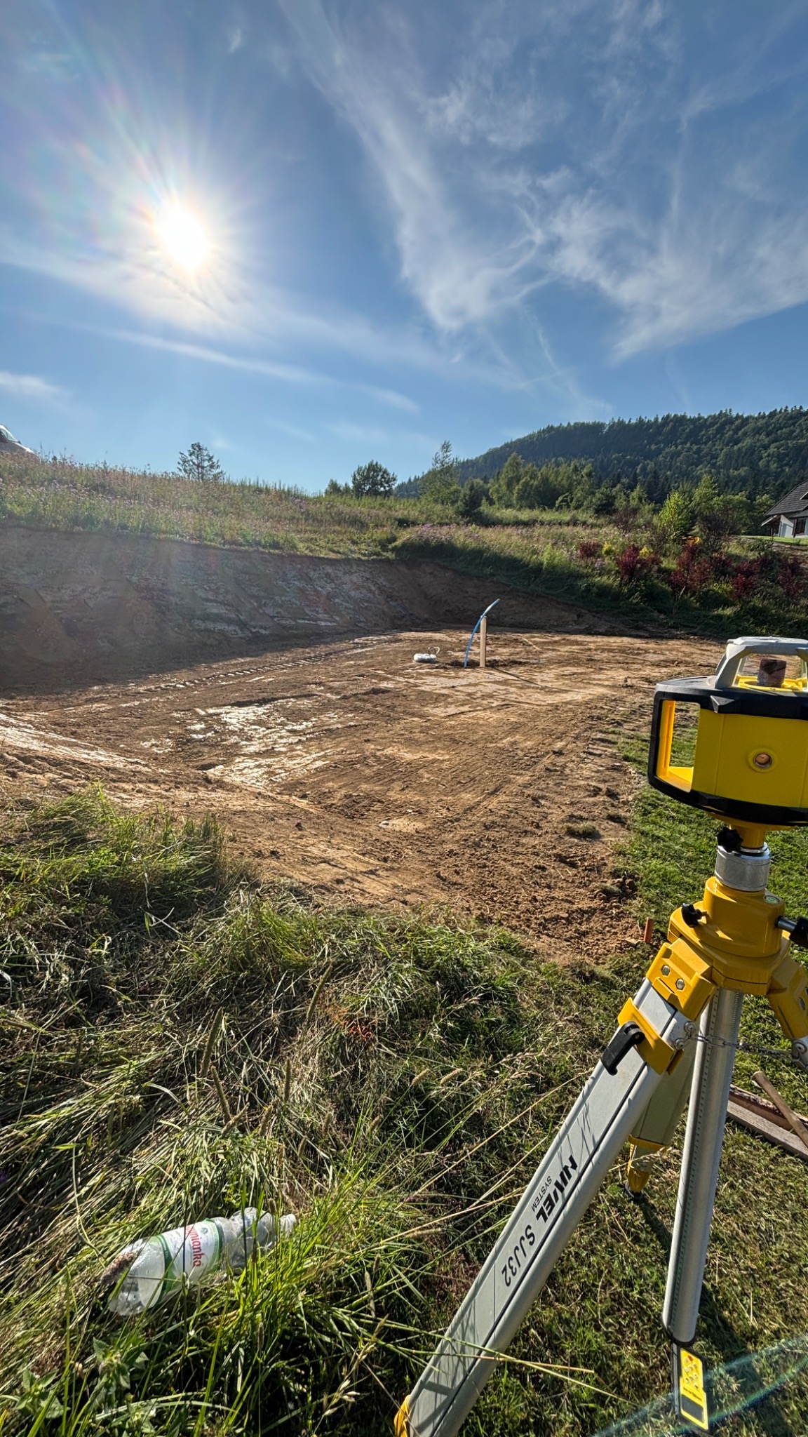Przygotowany teren pod budowę z widocznym niwelatorem laserowym na statywie. W tle zieleń i błękitne niebo z promieniami słońca. Butelka wody w trawie.