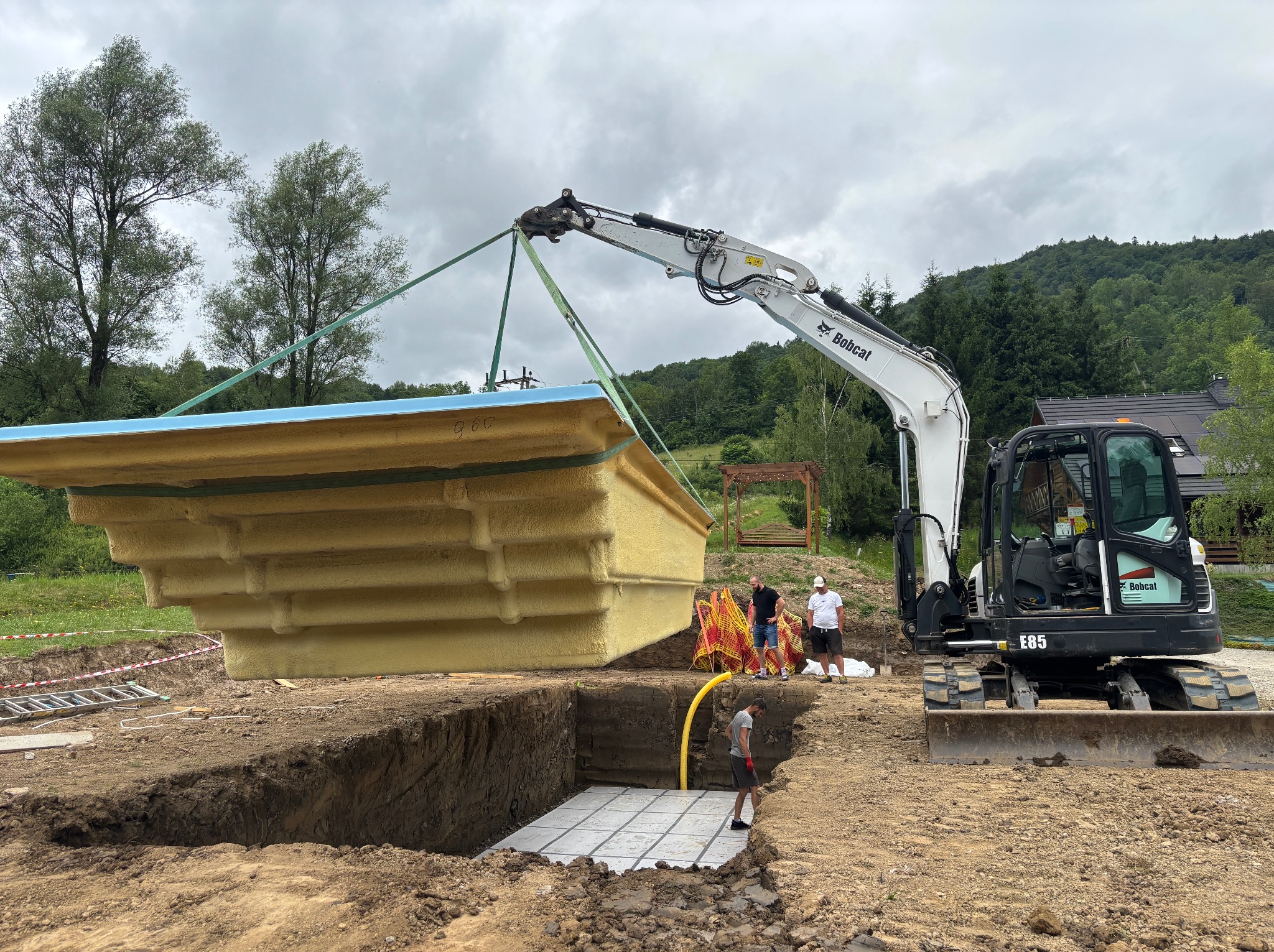 Bobcat E85 umieszcza prefabrykowany basen w wykopie na przygotowanym podłożu. W tle pracownicy i zalesione wzgórza.
