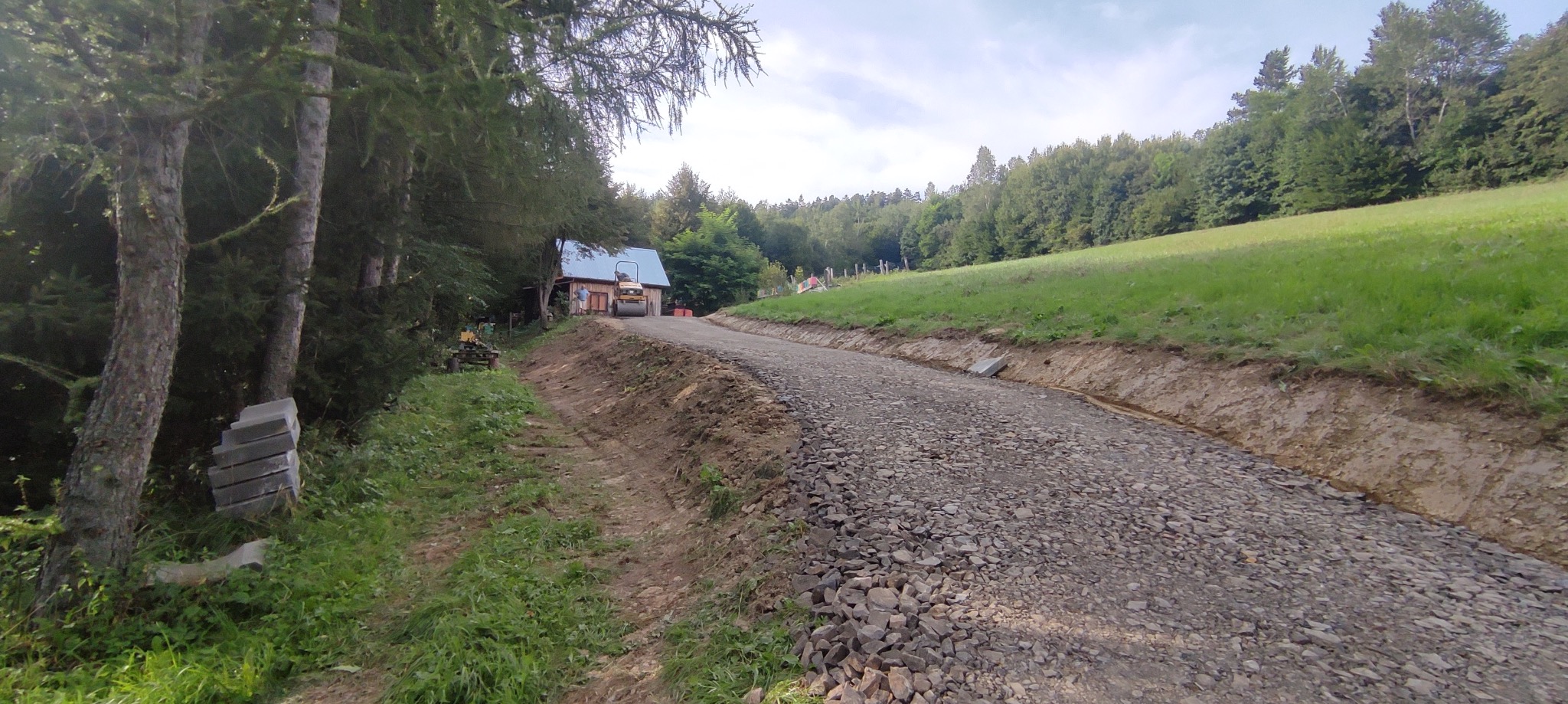 Utwardzona, szutrowa droga na zboczu, z widocznym walcem drogowym w oddali i stertą kostki brukowej przy drzewach. Zielone wzgórza i las w tle.