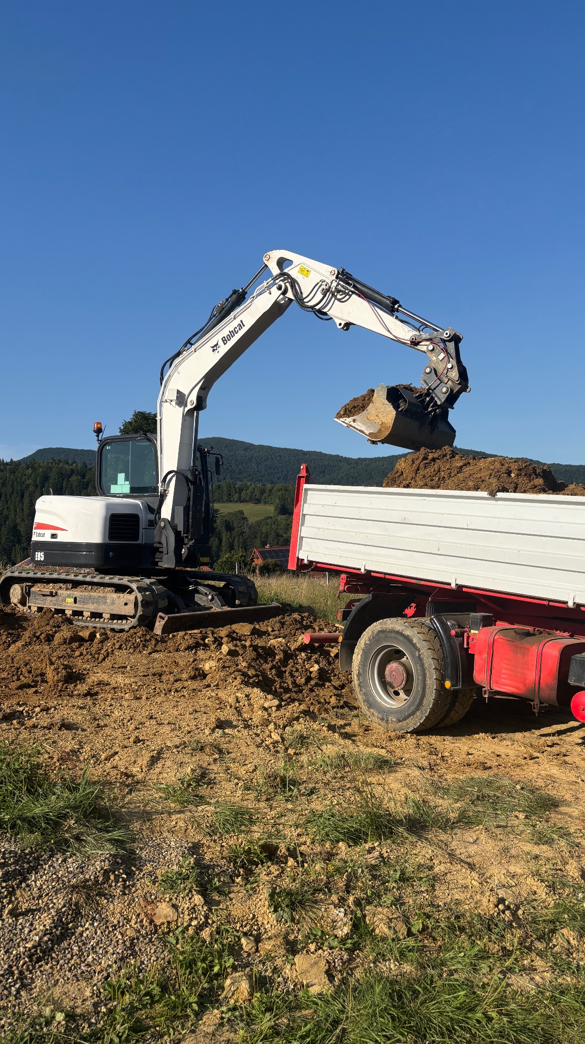 Biała koparka Bobcat E85 załadowuje ziemię do białej ciężarówki na tle zielonych wzgórz i błękitnego nieba. Widoczne ślady pracy ziemnej.