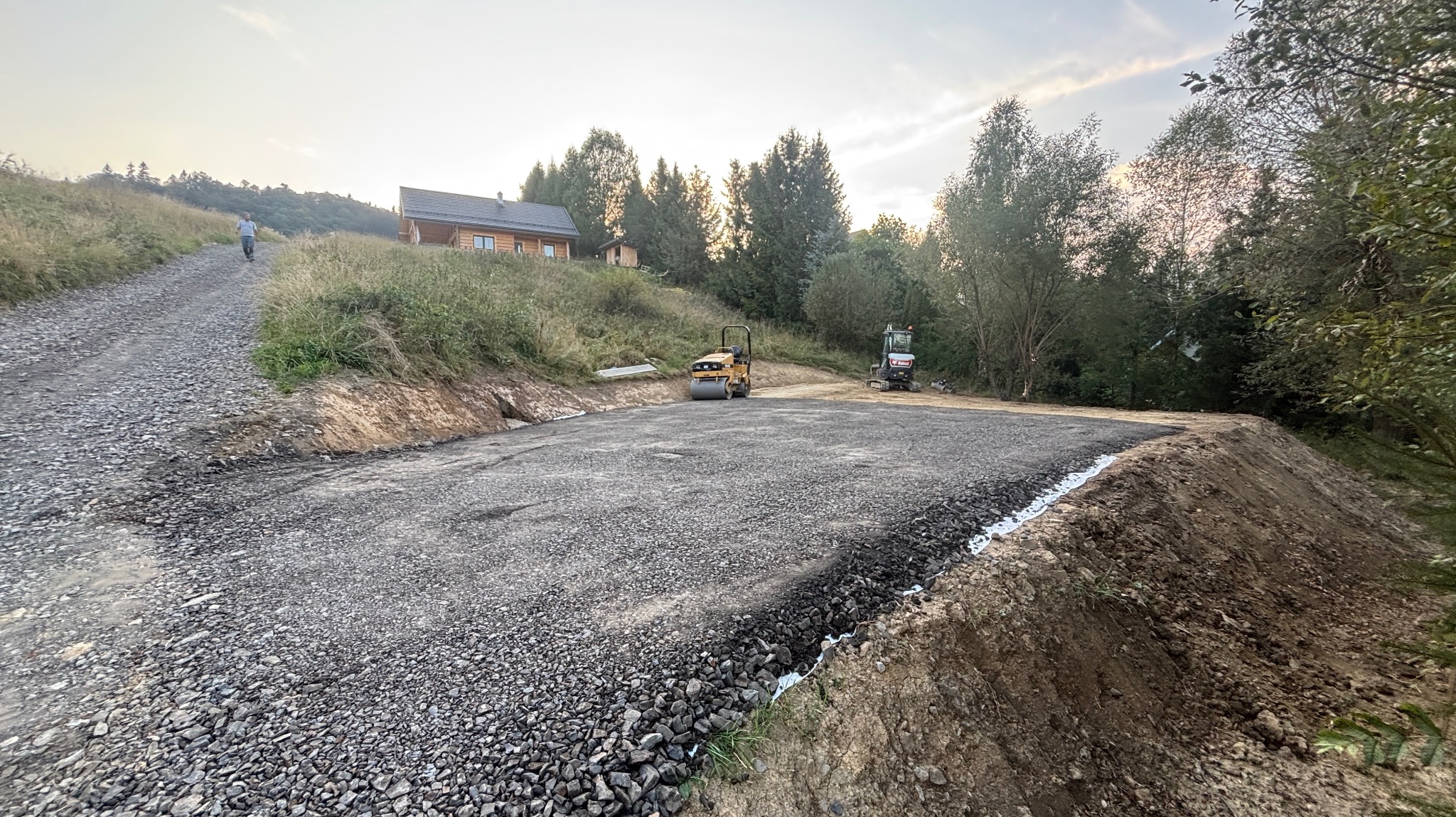Utwardzony teren pod budowę w górskim krajobrazie. Walec drogowy i minikoparka na przygotowanym placu. W tle drewniany dom i zalesione wzgórza.