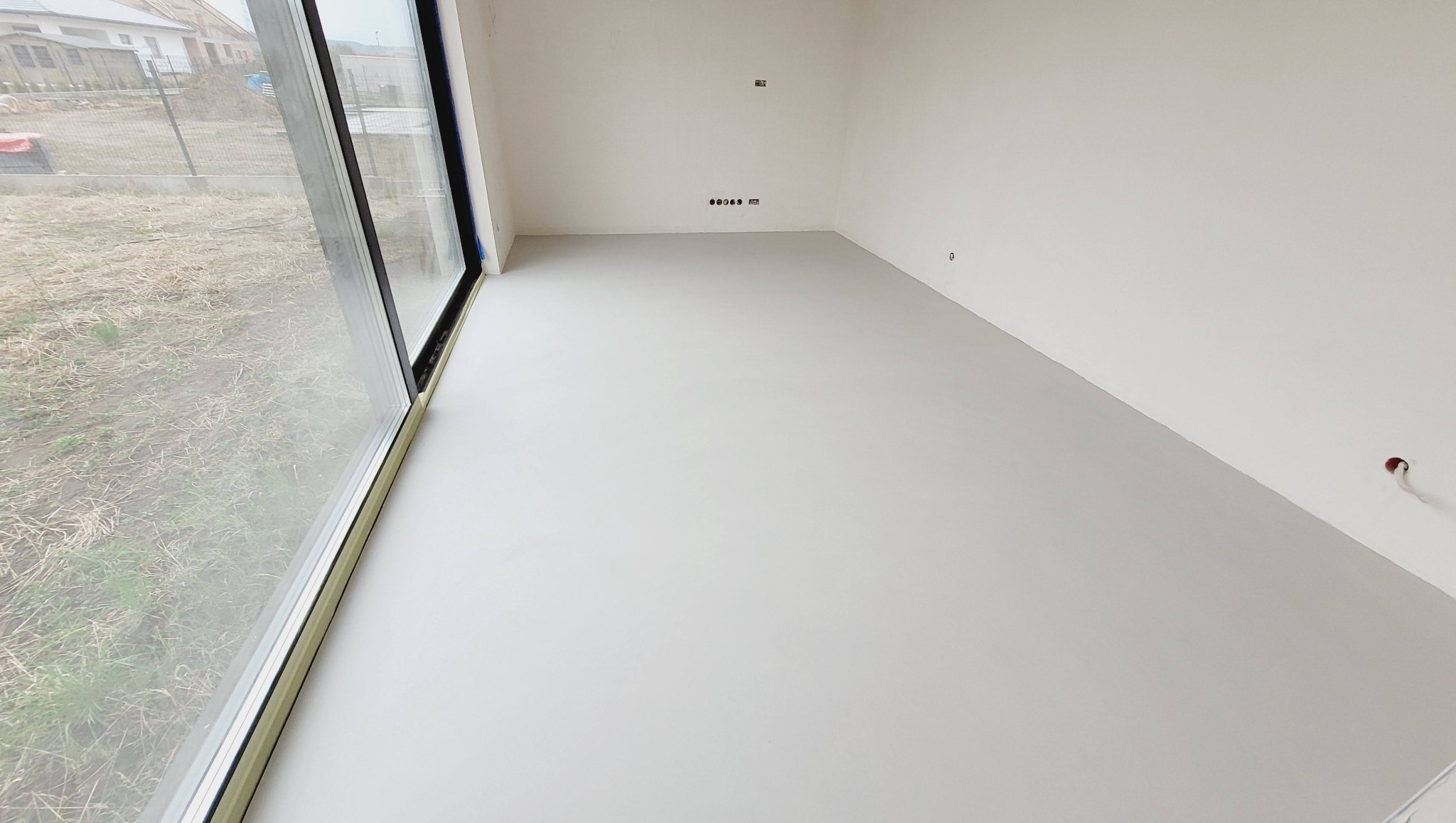 Jasne wnętrze z gładką, szarą posadzką i dużym oknem. Widoczne puszki elektryczne w ścianie. Minimalistyczny design, świeżo wykończone pomieszczenie.