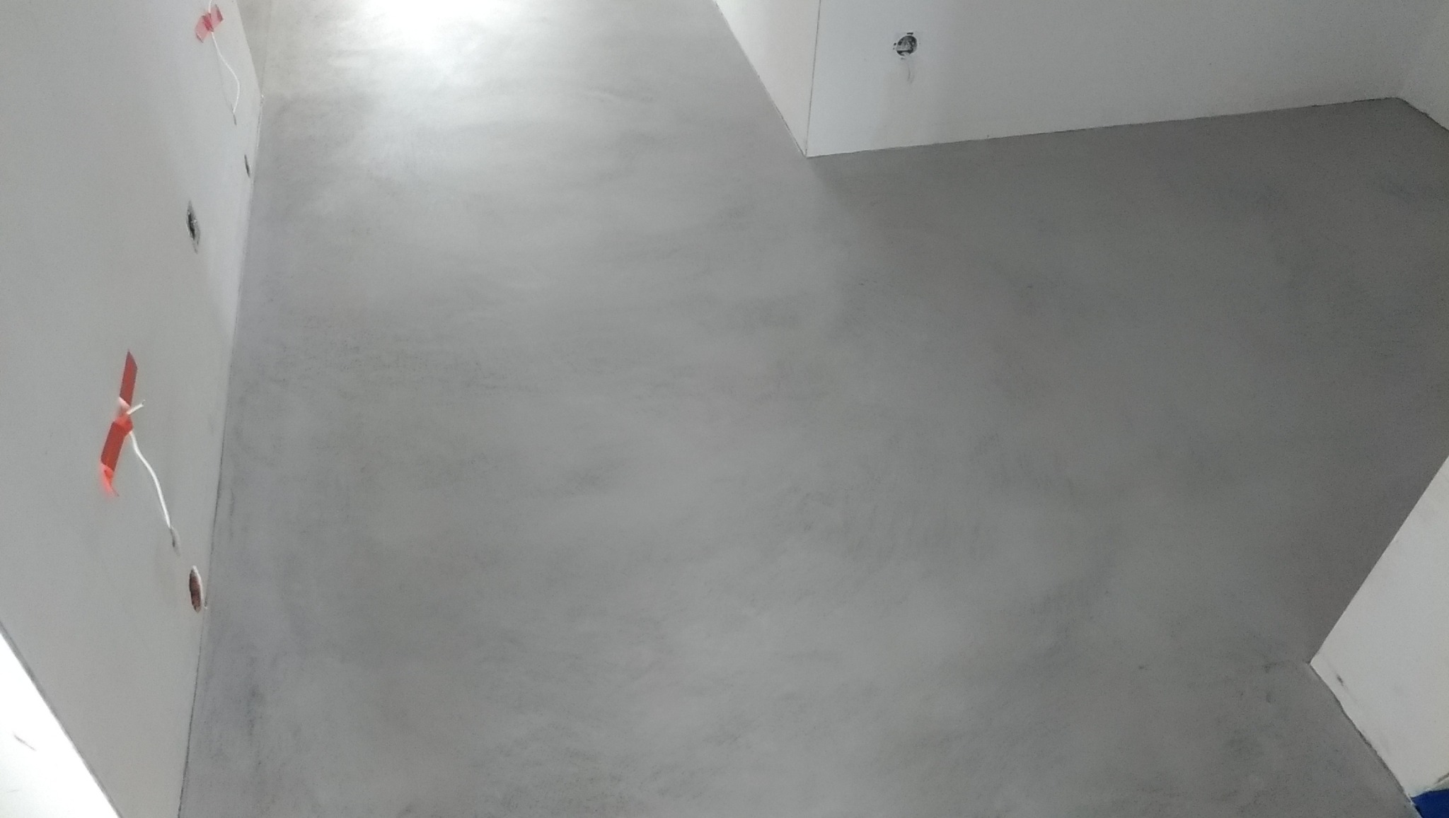 Szara, gładka posadzka w nowym wnętrzu, widoczne przygotowane punkty elektryczne na ścianie. Minimalistyczny design, surowy betonowy wygląd podłogi.