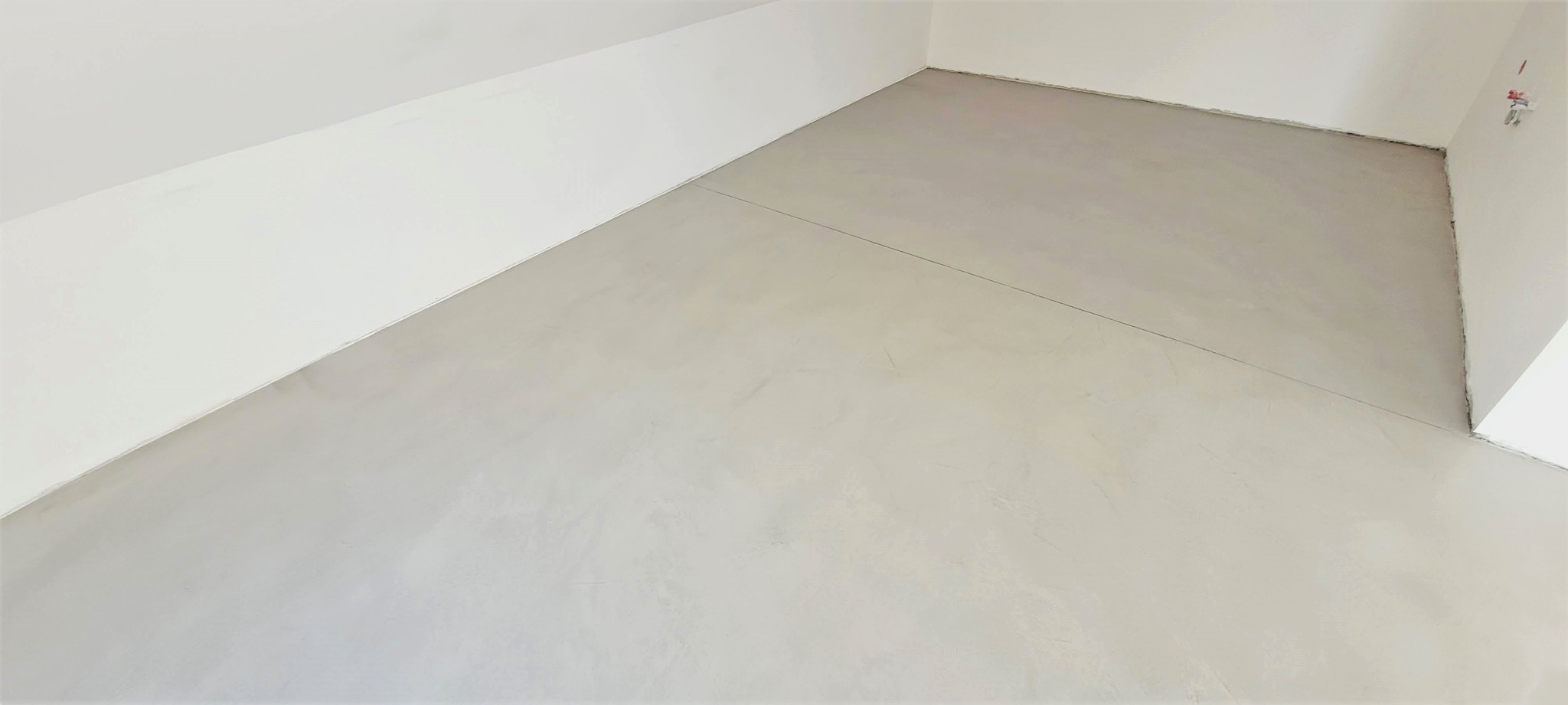 Jasnoszara, gładka posadzka w rogu pomieszczenia z białymi ścianami i skośnym sufitem. Minimalistyczne wykończenie wnętrza, widok z góry.