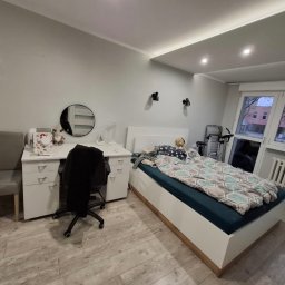 Jakub Jędrosek INTERIORS Twój styl Nasze wykończenia - Jasne wnętrze pokoju z łóżkiem, biurkiem i drewnianą podłogą. Ściany pomalowane na szaro, sufit z oświetleniem LED. Widoczne okno z grzejnikiem.