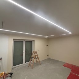 Jakub Jędrosek INTERIORS Twój styl Nasze wykończenia - Pokój po remoncie z zamontowanym oświetleniem LED na suficie. Widoczna drabina, narzędzia malarskie i częściowo pomalowane ściany. Podłoga szara, surowa.