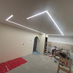 Jakub Jędrosek INTERIORS Twój styl Nasze wykończenia - Wnętrze w trakcie remontu: gładkie ściany, sufit z geometrycznym oświetleniem LED, narzędzia malarskie i drabina. Widoczny fragment podłogi zabezpieczony folią.