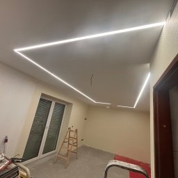 Jakub Jędrosek INTERIORS Twój styl Nasze wykończenia - Pokój w trakcie remontu z zamontowanym oświetleniem LED na suficie, drabina oparta o okno, widoczne narzędzia malarskie i niedokończone ściany.