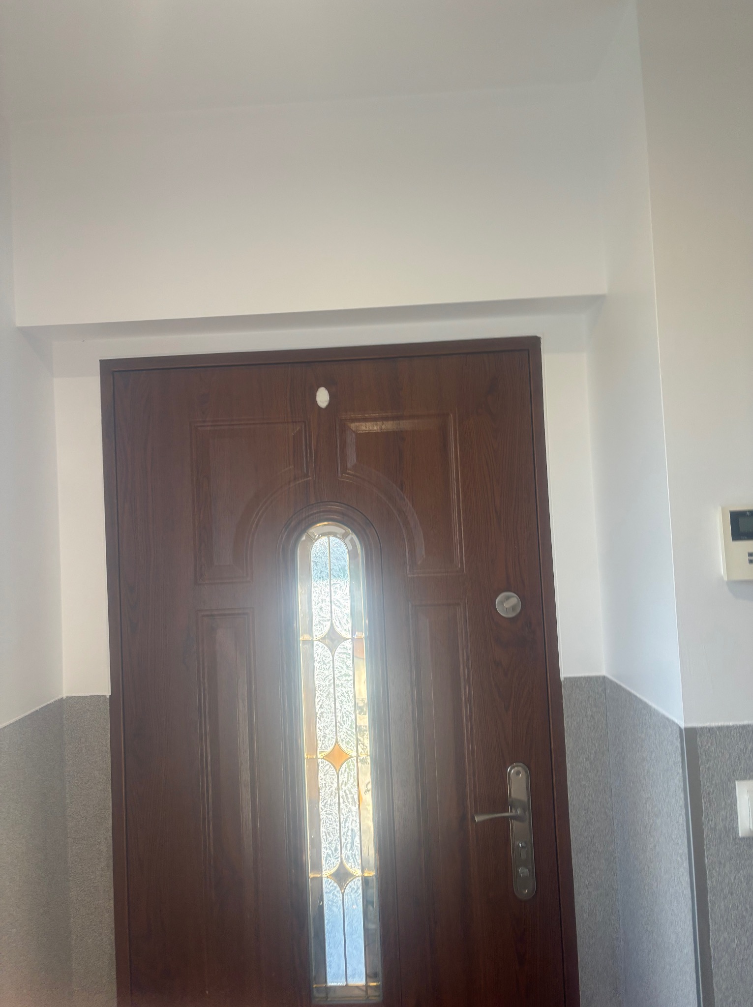 Ciemnobrązowe drzwi wejściowe z szybą witrażową, osadzone w świeżo pomalowanej na biało ścianie z szarą lamperią. Widoczne detale wykończenia.