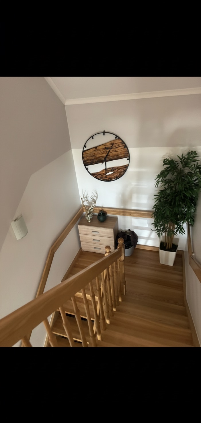 Schody z drewnianą balustradą, na ścianie zegar z drewnianym elementem i żywicy, komoda z dekoracjami, roślina w doniczce. Widok z góry.