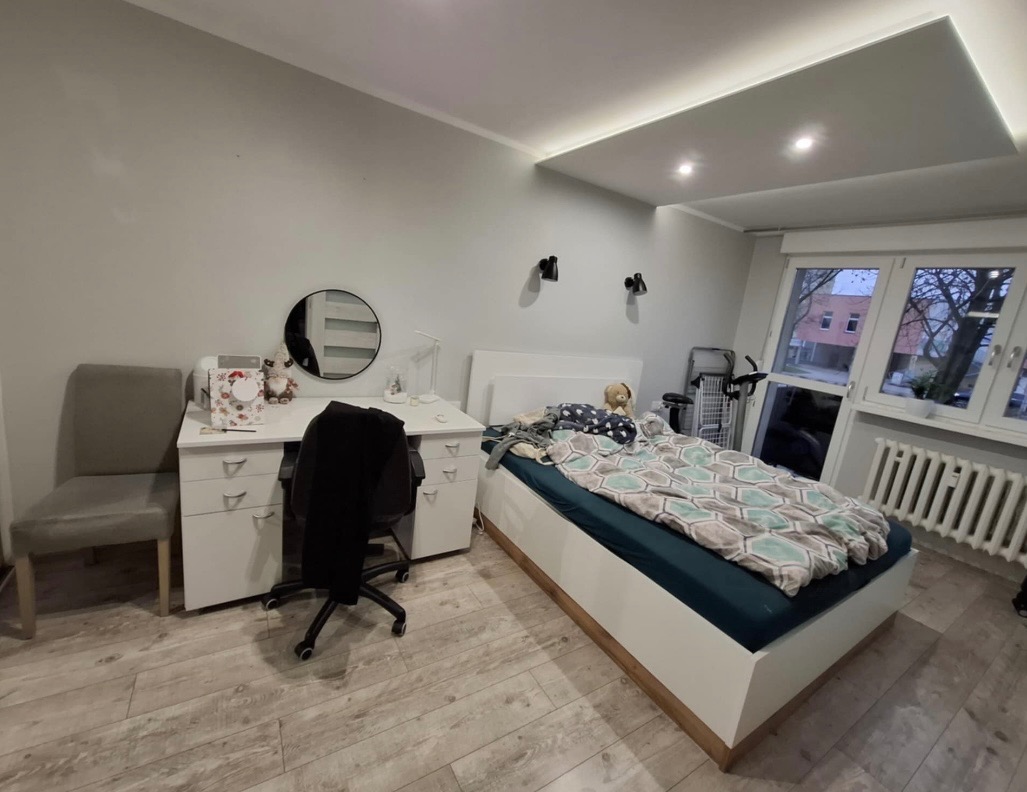 Jasne wnętrze pokoju z łóżkiem, biurkiem i drewnianą podłogą. Ściany pomalowane na szaro, sufit z oświetleniem LED. Widoczne okno z grzejnikiem.