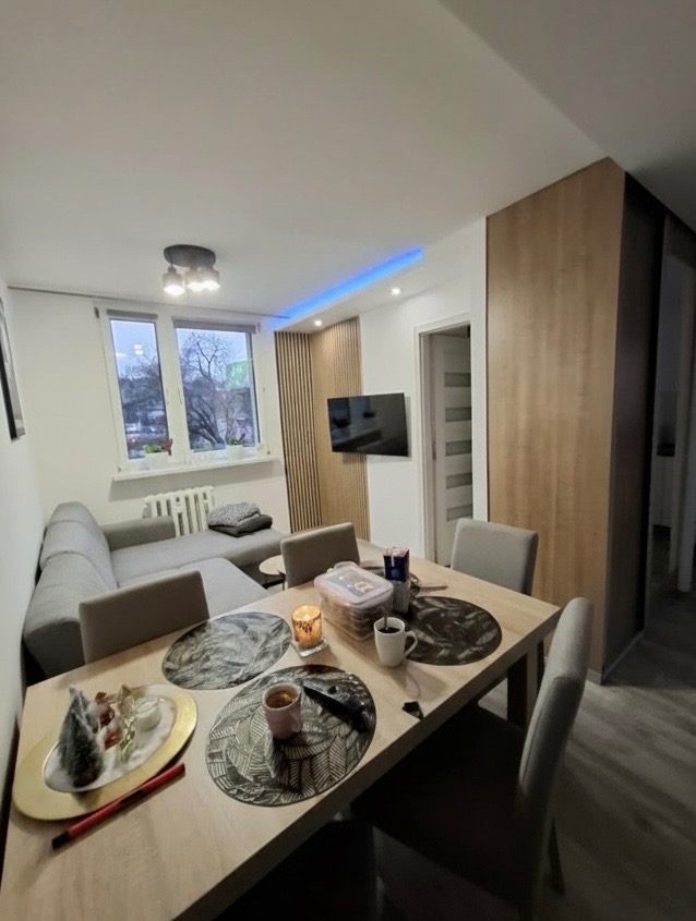 Jasne wnętrze po remoncie: drewniane panele ścienne, szara sofa narożna, stół z dekoracjami. Nowoczesne oświetlenie LED podkreśla aranżację.