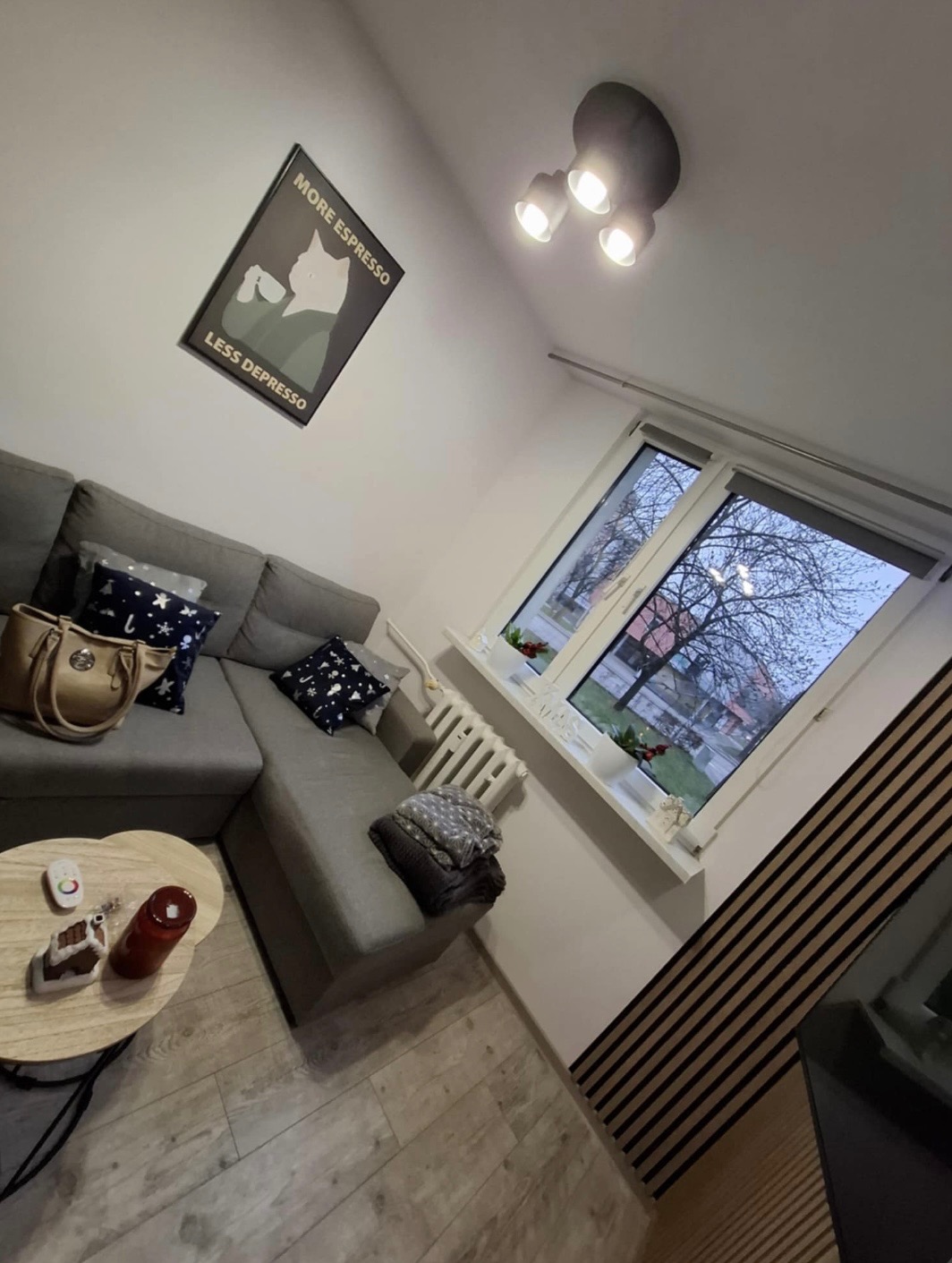 Przytulny salon z szarą sofą narożną, drewnianą ścianą z lameli i plakatem 'More Espresso Less Depresso'. Widok na okno z parapetem i grzejnikiem.