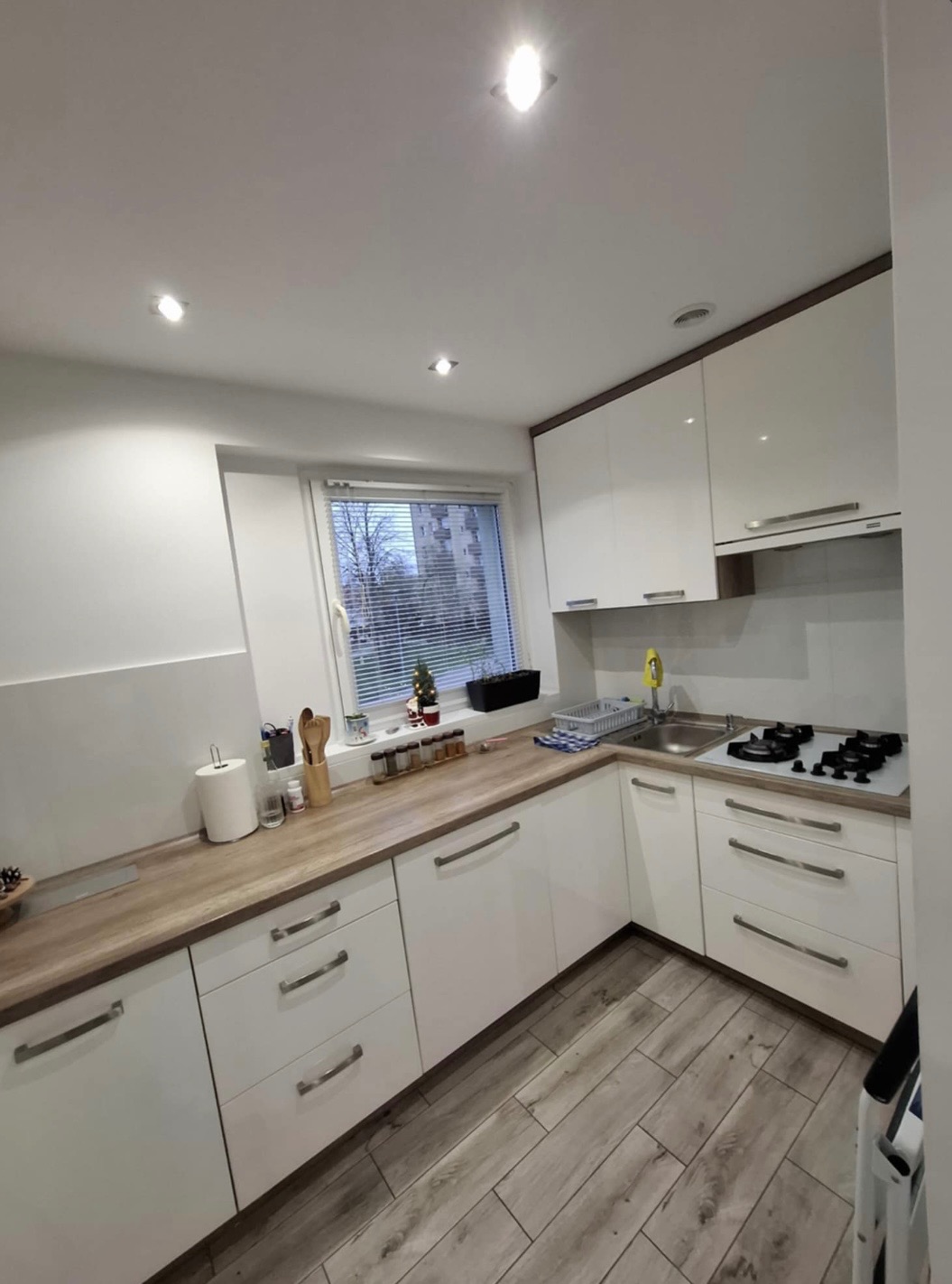 Jasna kuchnia w stylu skandynawskim z drewnianym blatem i białymi frontami szafek. Widoczne okno z żaluzjami i drewniana podłoga imitująca panele.