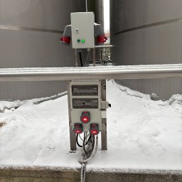 Szara skrzynka elektryczna z gniazdami i przewodami na tle zbiorników przemysłowych pokrytych śniegiem. Widoczne złącza siłowe i zabezpieczenia.
