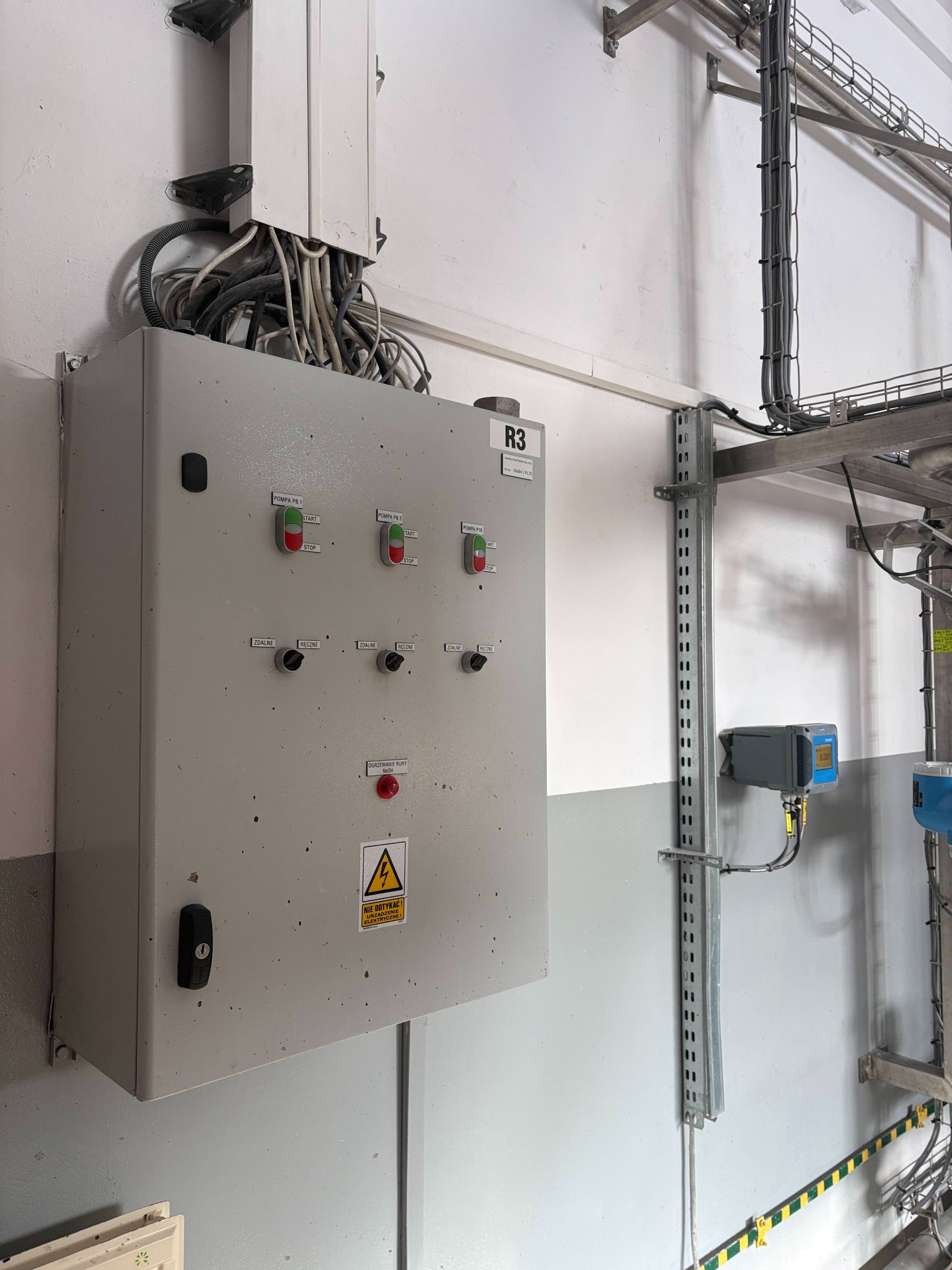 Szara szafa sterownicza z przyciskami i oznaczeniami, zamontowana na ścianie w pomieszczeniu przemysłowym. Widoczne kable i instalacje elektryczne.