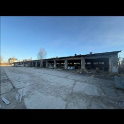 Ekspres Multiserwis - Długi, parterowy budynek w stanie rozkładu z otwartymi wjazdami, otoczony betonowym placem i pojedynczym drzewem. Widok w słoneczny dzień.