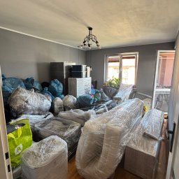 Ekspres Multiserwis - Pokój przed przeprowadzką: meble i przedmioty zabezpieczone folią, worki z rzeczami, gotowe do transportu. Widok na okno i drzwi.