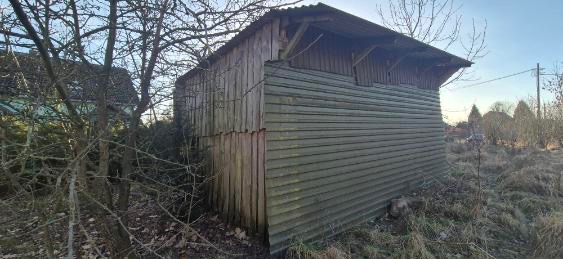Stary, drewniany budynek gospodarczy z blachy falistej na dachu, otoczony suchą trawą i drzewami bez liści, w tle widoczny dom.