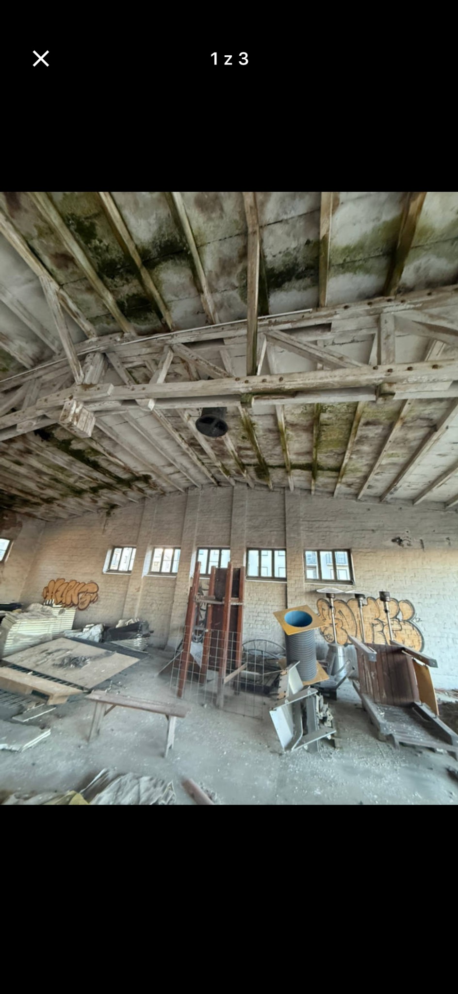 Wnętrze opuszczonego magazynu z graffiti na ścianach, drewnianą konstrukcją dachu i porozrzucanymi elementami wyposażenia. Widok z dołu, surowy klimat.