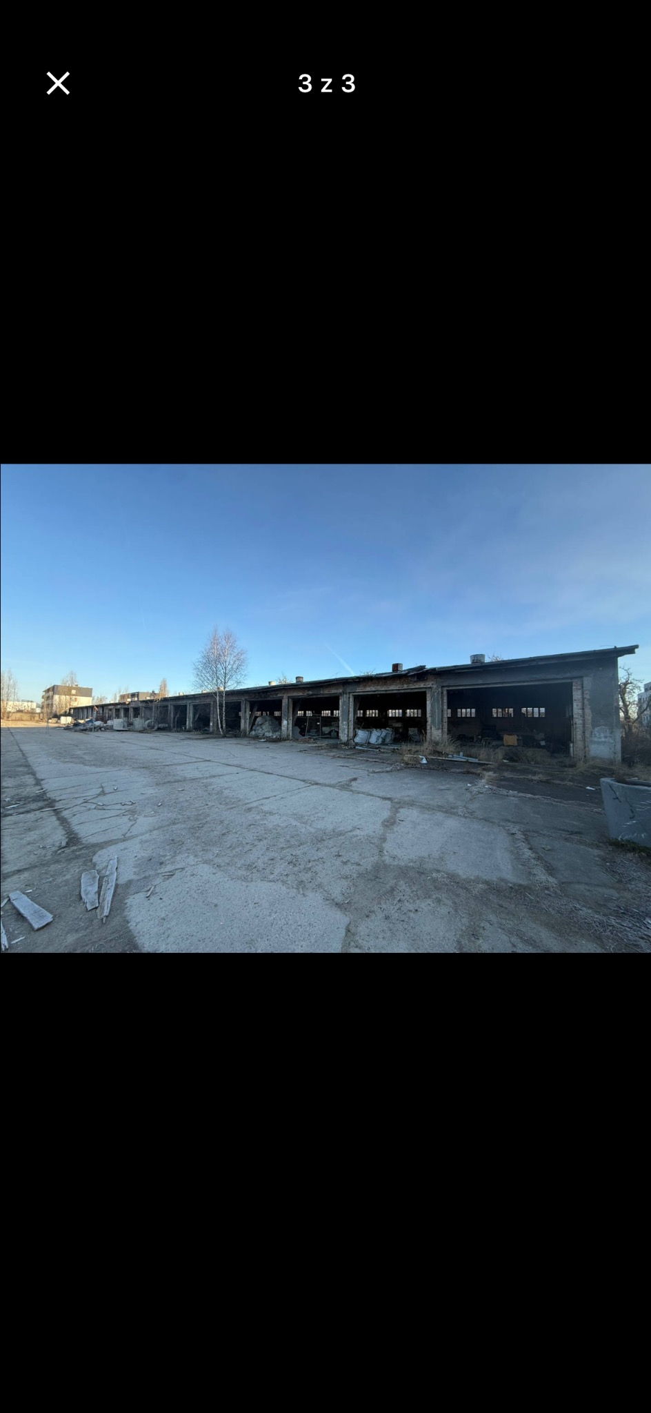 Długi, parterowy budynek w stanie rozkładu z otwartymi wjazdami, otoczony betonowym placem i pojedynczym drzewem. Widok w słoneczny dzień.