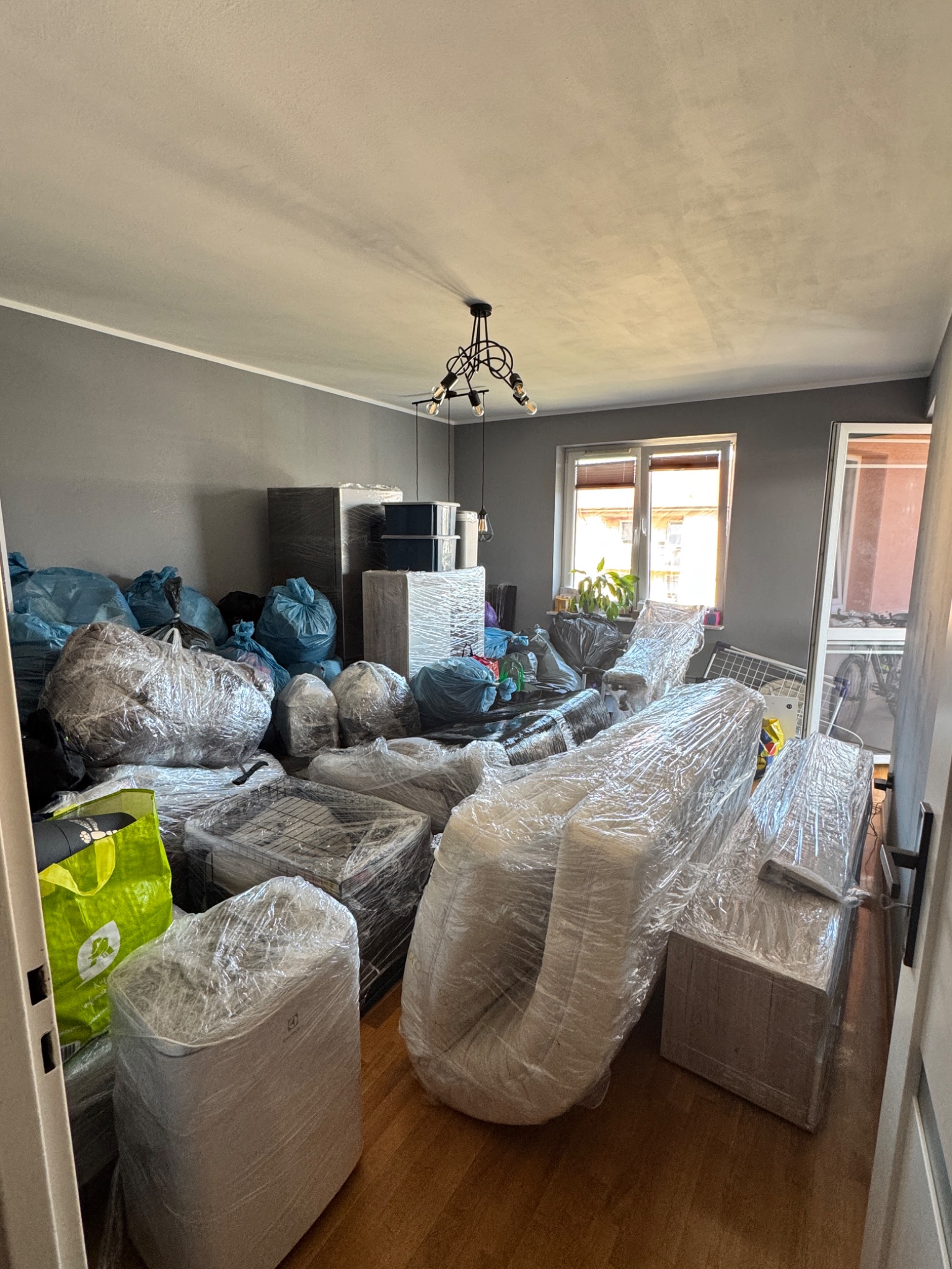 Pokój przed przeprowadzką: meble i przedmioty zabezpieczone folią, worki z rzeczami, gotowe do transportu. Widok na okno i drzwi.