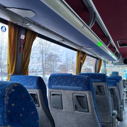 Przewozy „Yellon Bus”' Sławomir Jelonek - Wnętrze autobusu z rzędem niebiesko-szarych foteli pasażerskich, widok na okno z żółtą zasłoną i uchwytem do młotka bezpieczeństwa.