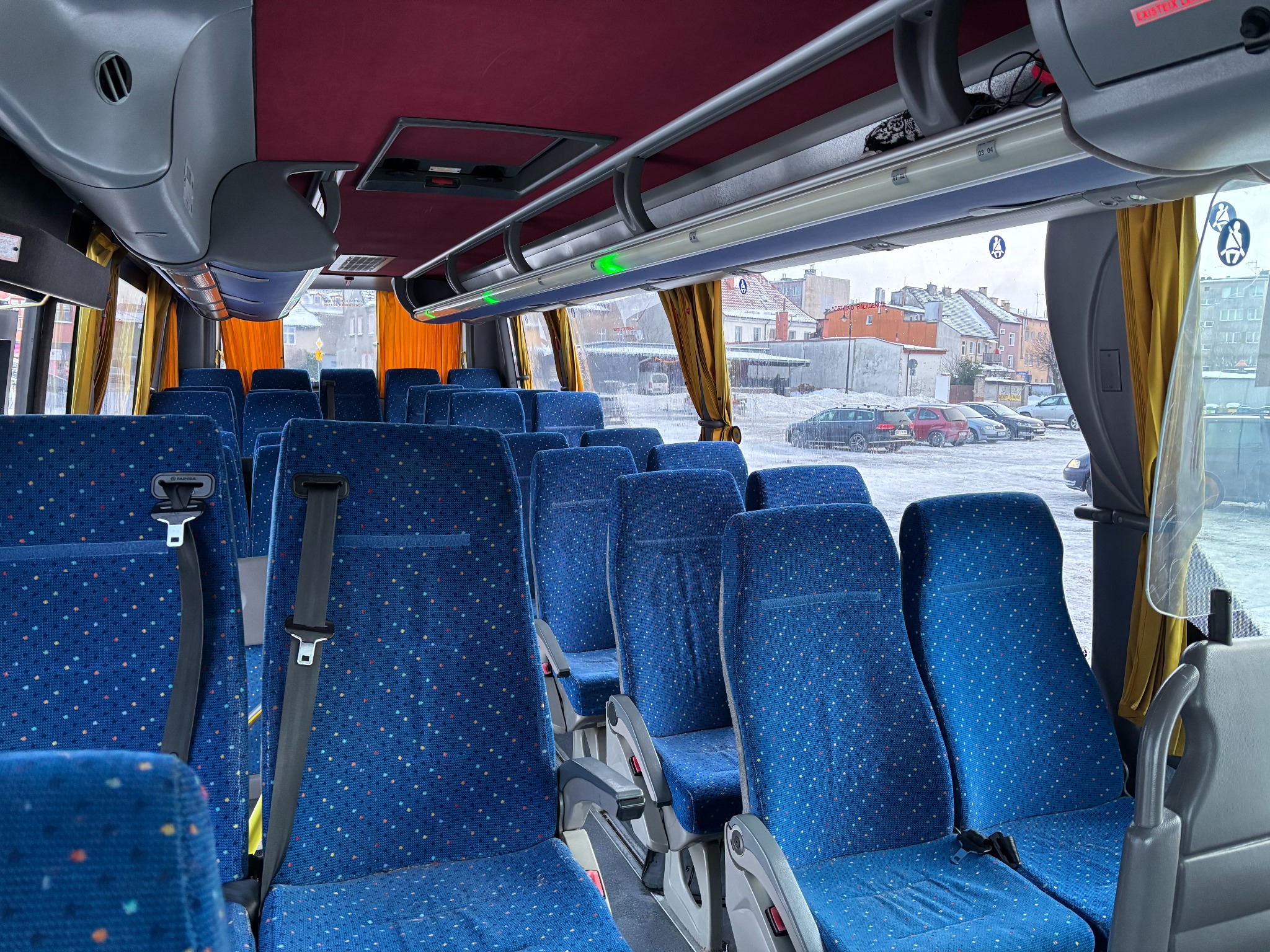 Wnętrze autobusu z niebieskimi siedzeniami w kropki, widok na okno z zimowym krajobrazem. Żółte zasłony i szare elementy wyposażenia.