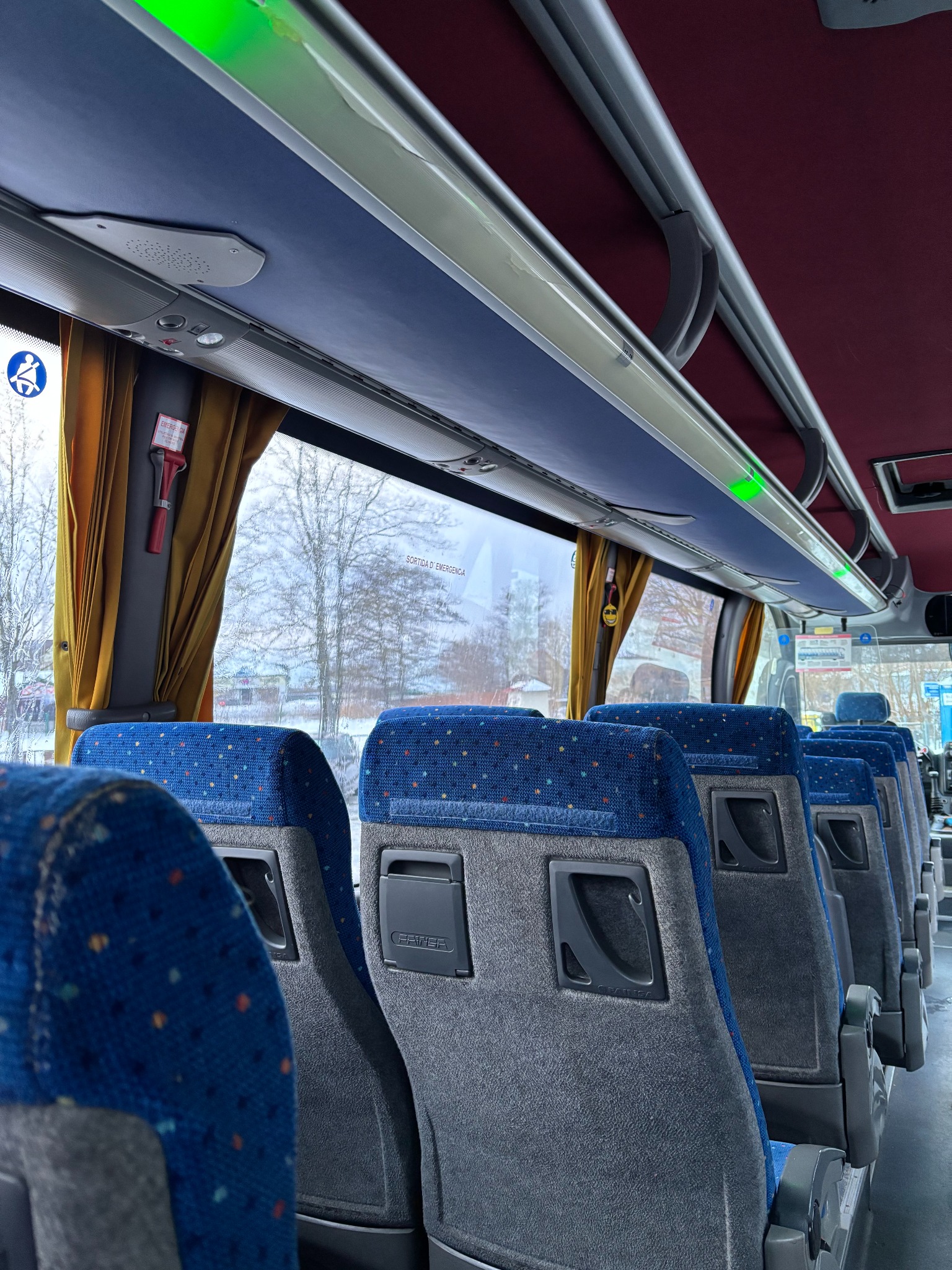 Wnętrze autobusu z rzędem niebiesko-szarych foteli pasażerskich, widok na okno z żółtą zasłoną i uchwytem do młotka bezpieczeństwa.